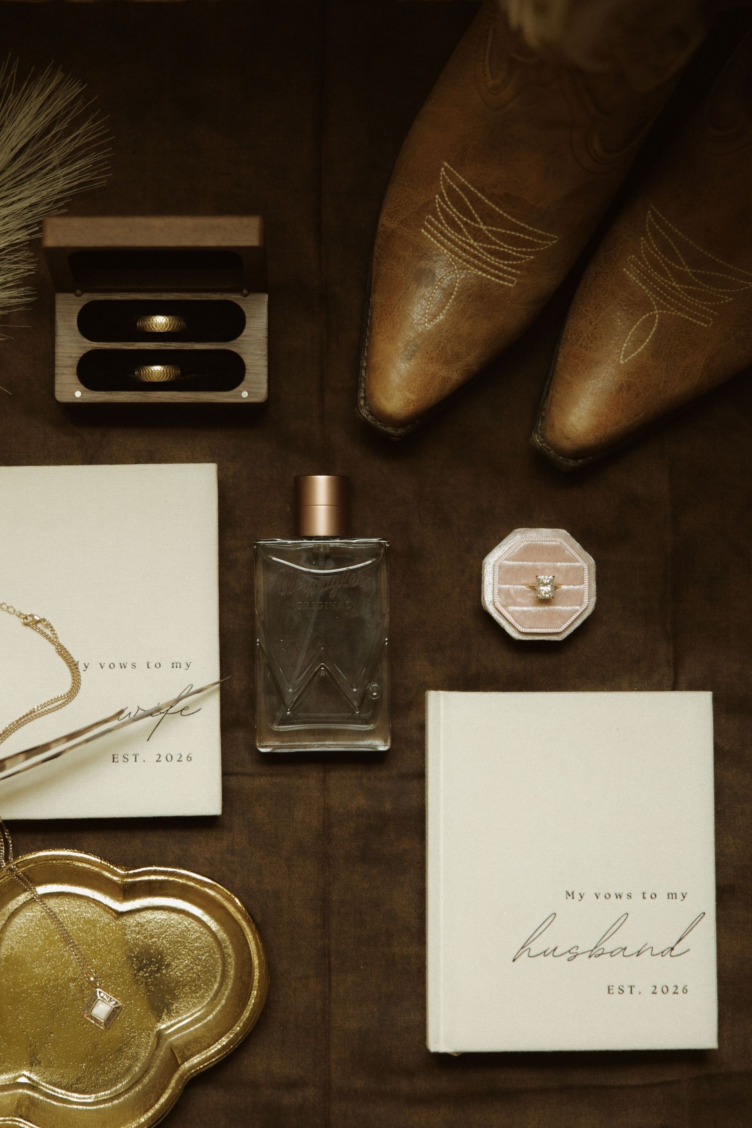 Rustic wedding flat lay with cowboy boots, vow books, perfume, ring box, and gold tray on dark wood surface.