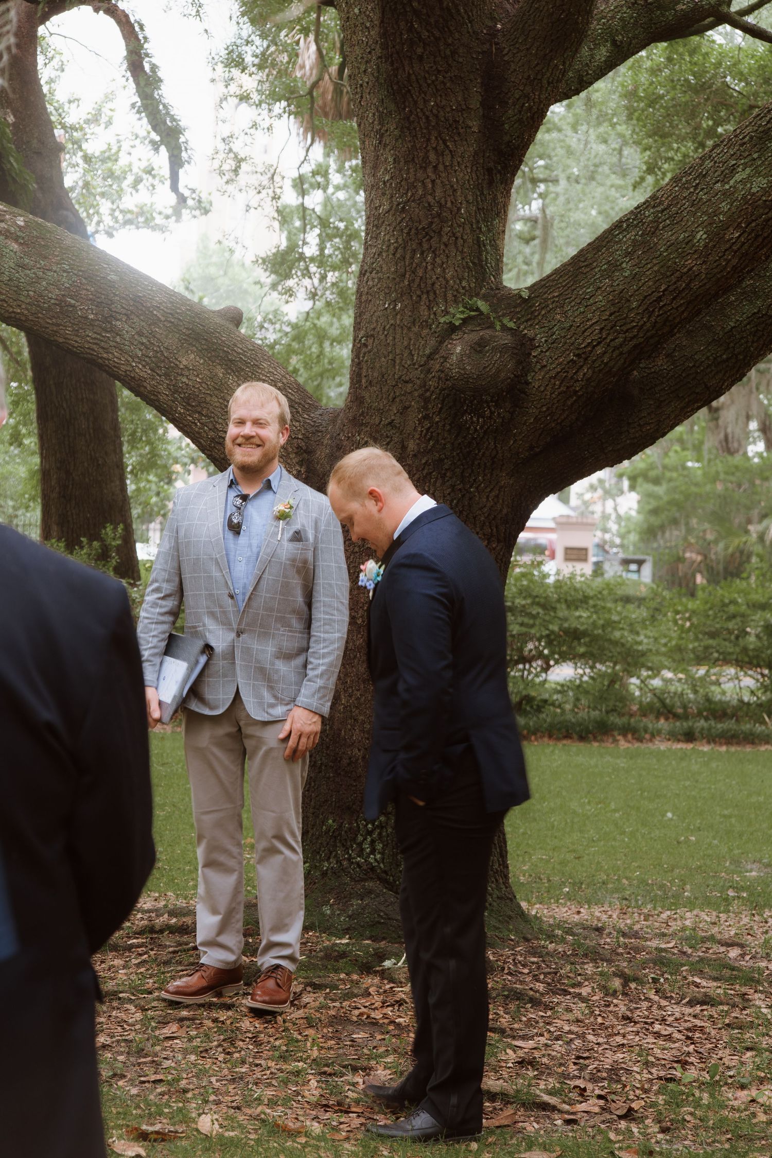 Two men in suits stand beneath the spreading branches of a large oak tree during what appears to be a wedding ceremony.