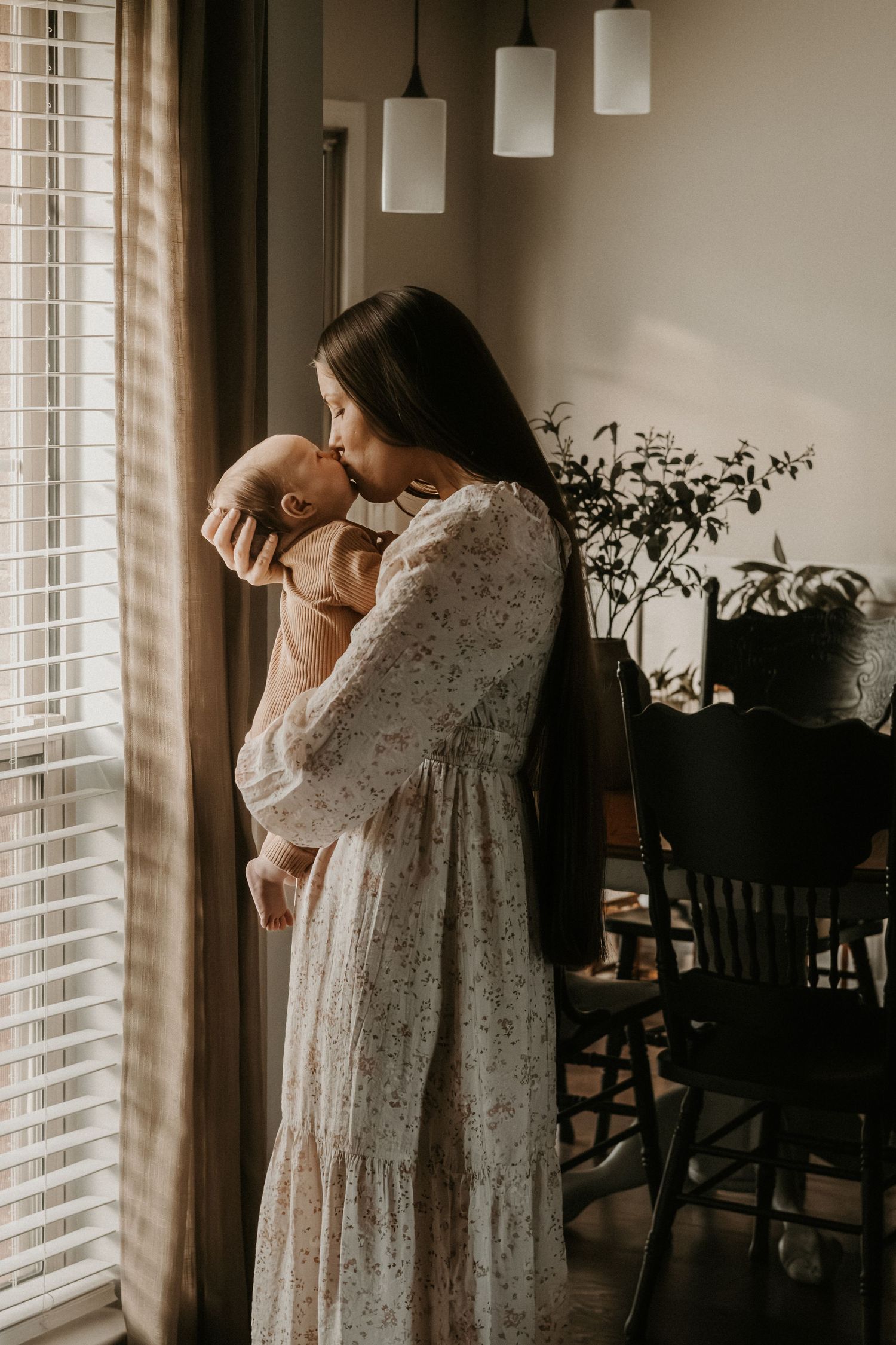 Tender moment by window light as parent cuddles newborn in flowing dress.