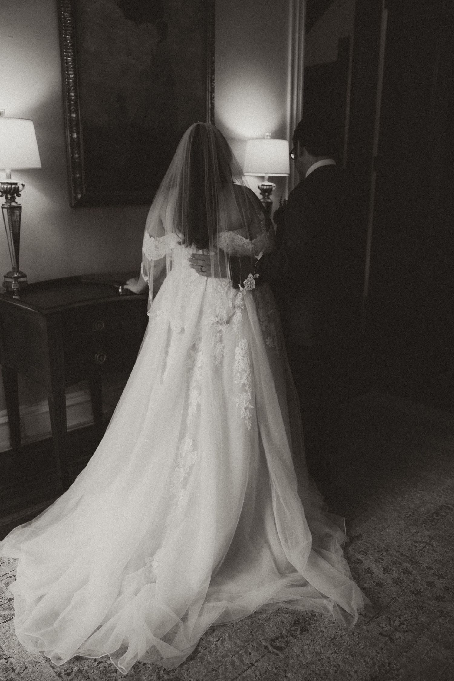 Black and white photograph of a bride in a flowing lace wedding gown with long veil standing indoors.