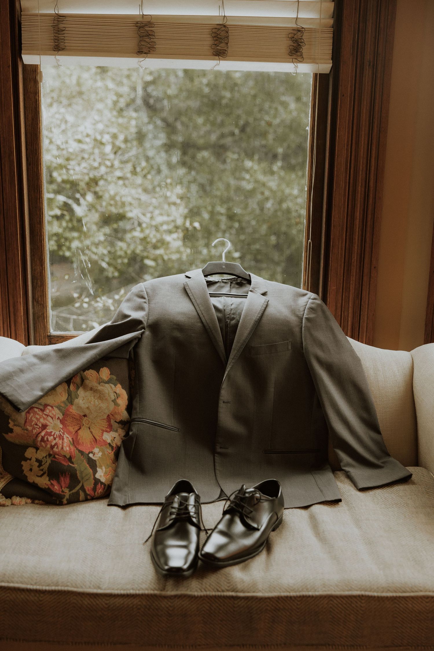 Gray suit jacket and dress shoes arranged on beige couch near window.