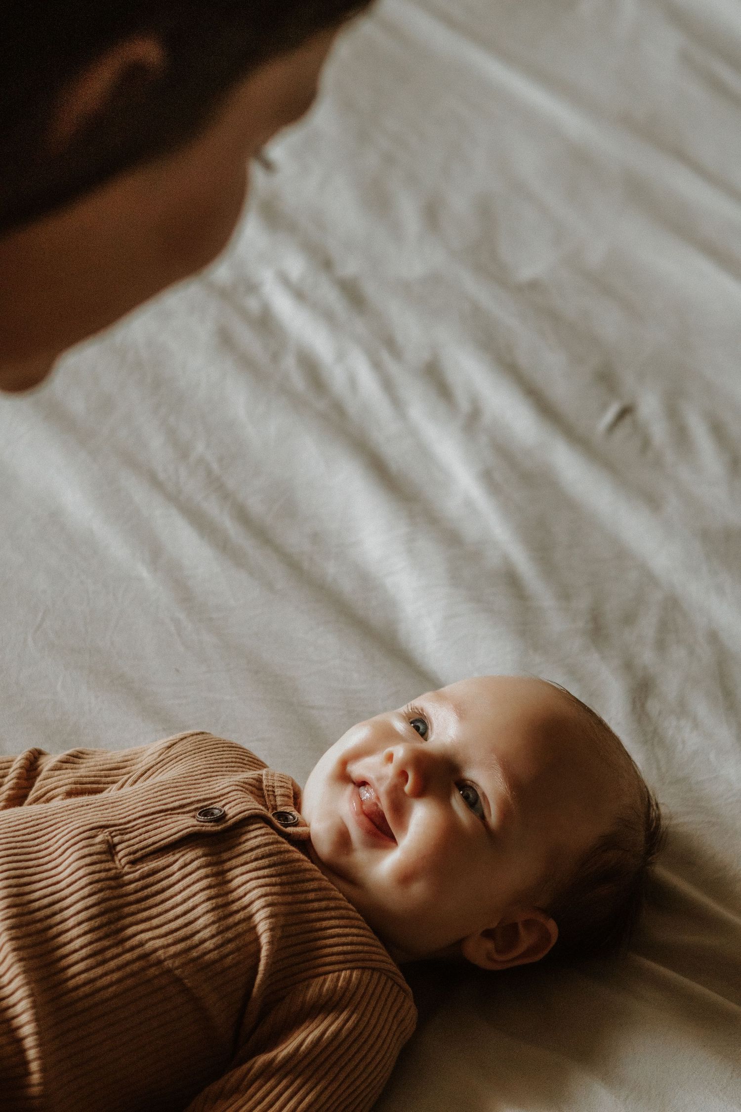 Baby in brown ribbed sweater smiling while lying on white bedding.