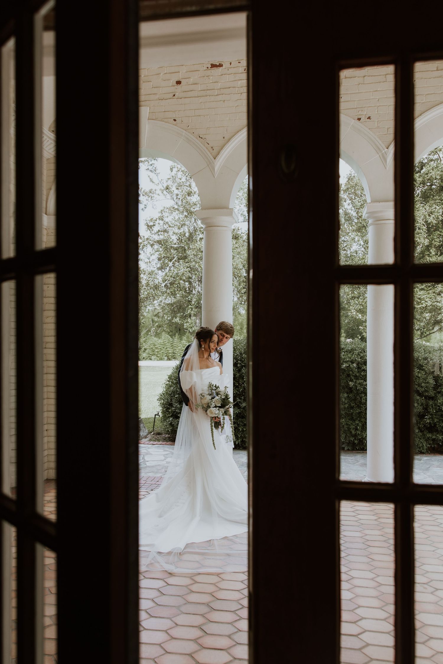 Wedding portrait through columns showing a bride in white dress on an elegant porch with classic architecture and greenery.