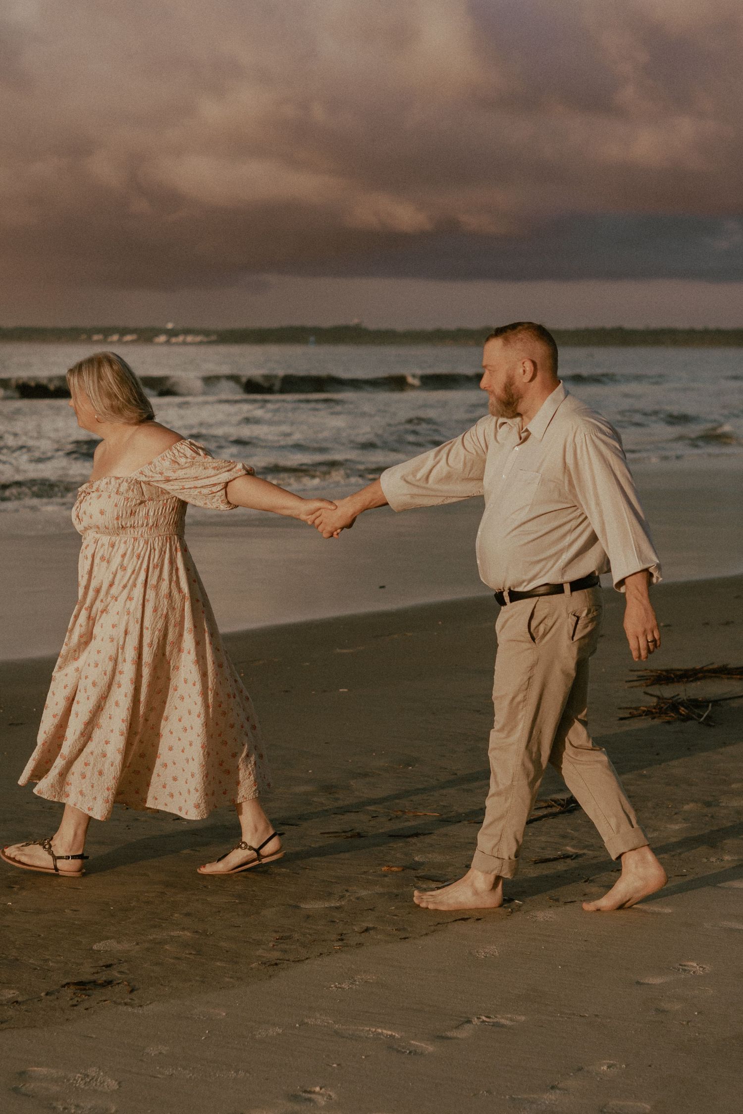 Couple holding hands and walking along a beach at sunset with dramatic cloudy skies.