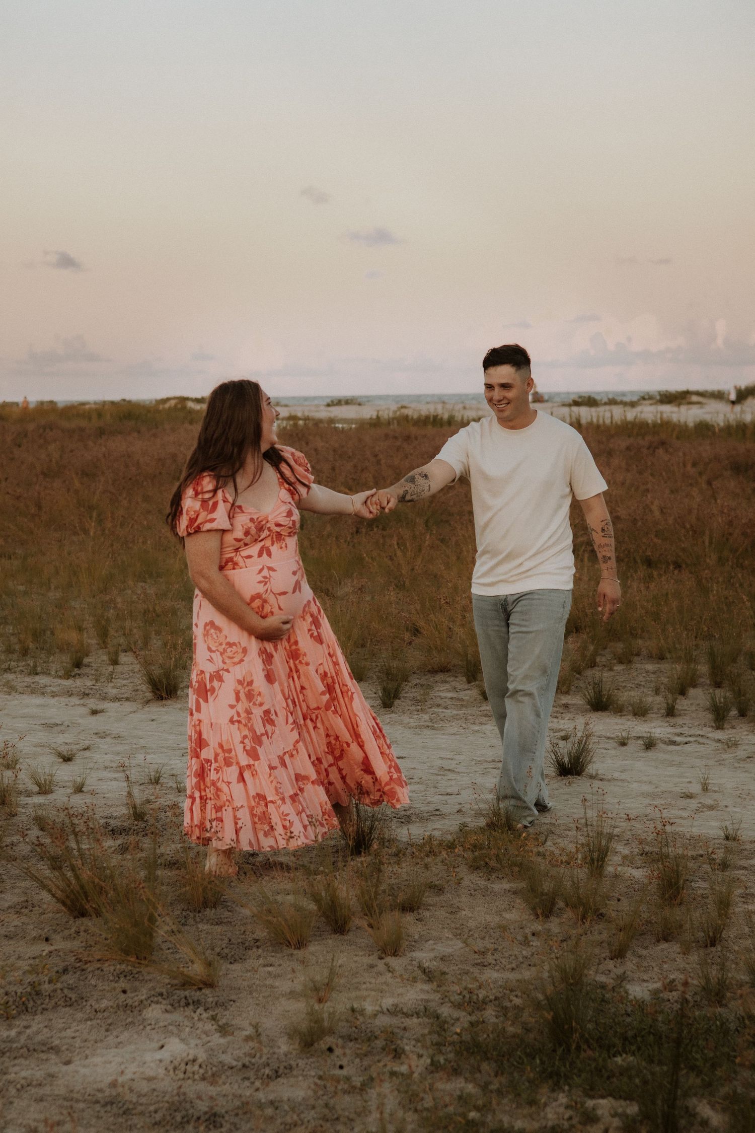 Couple holding hands while walking through coastal field at sunset.