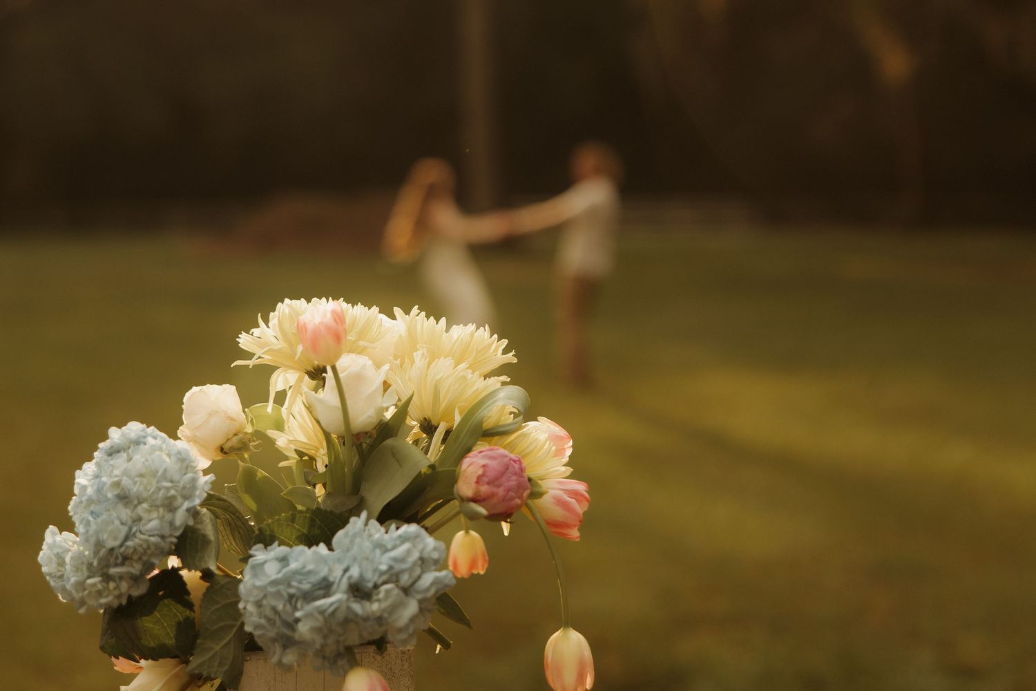 A vibrant bouquet of pink, white, and blue flowers in the foreground with two people dancing in a sunny green field.