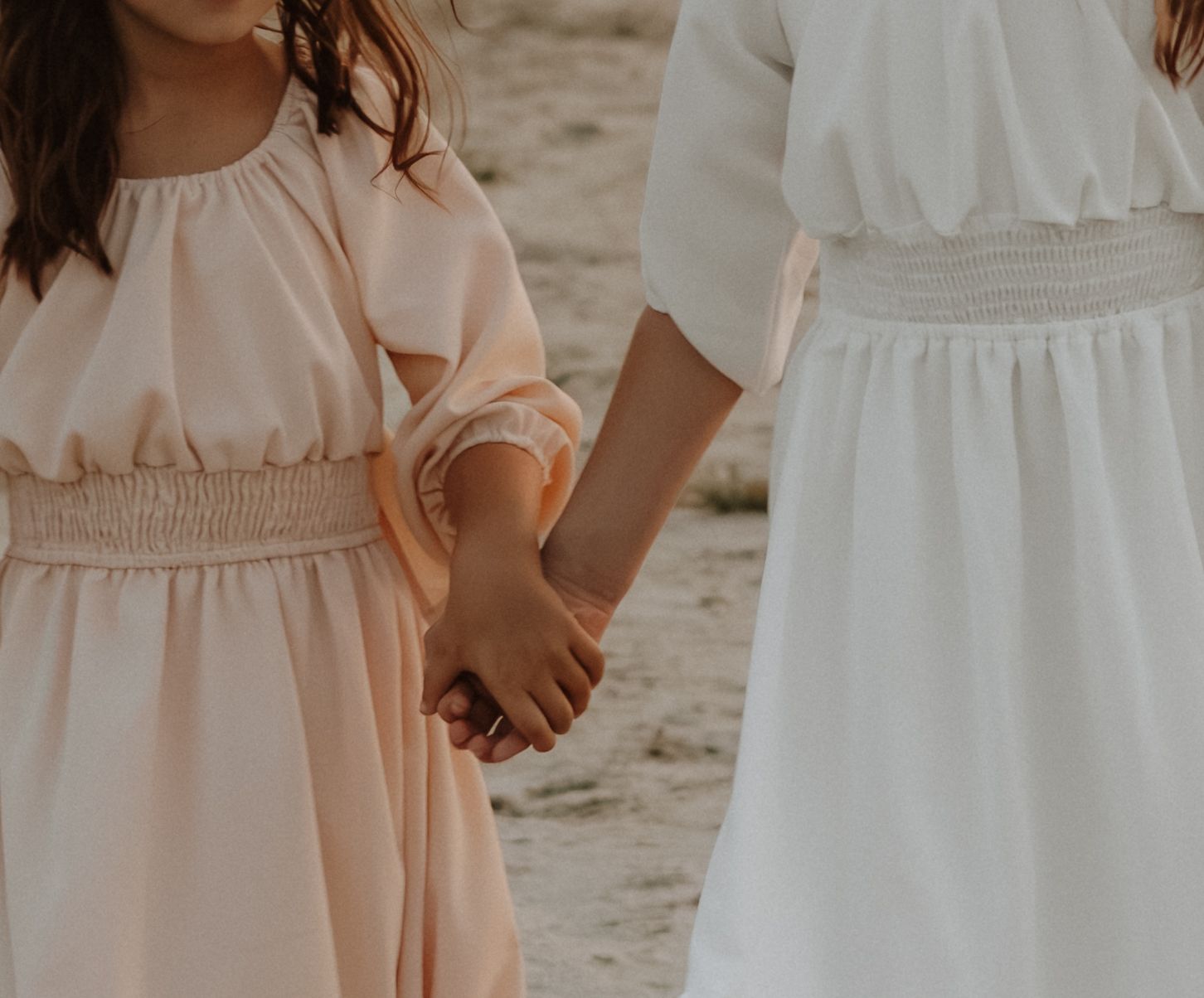 Two people in flowing white and blush dresses hold hands on a sandy beach at sunset.