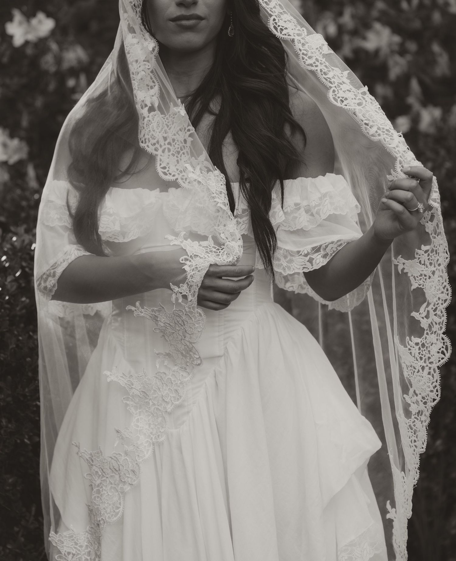 Black and white photo of a bride in a lace wedding dress and veil, holding her veil gracefully outdoors.