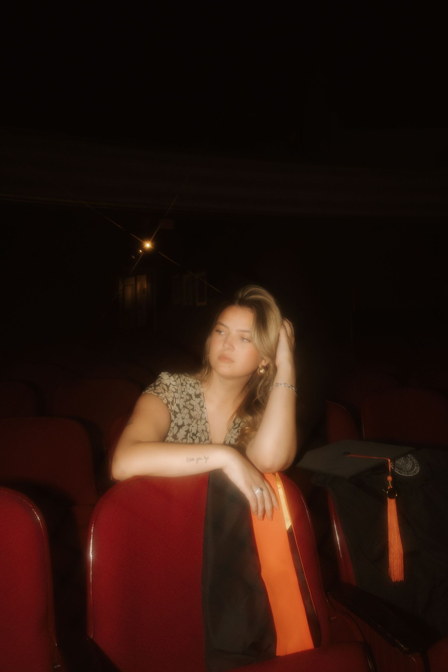 Young woman sitting alone in red theater seats, wearing a floral top, resting her head on her hand in dim light.