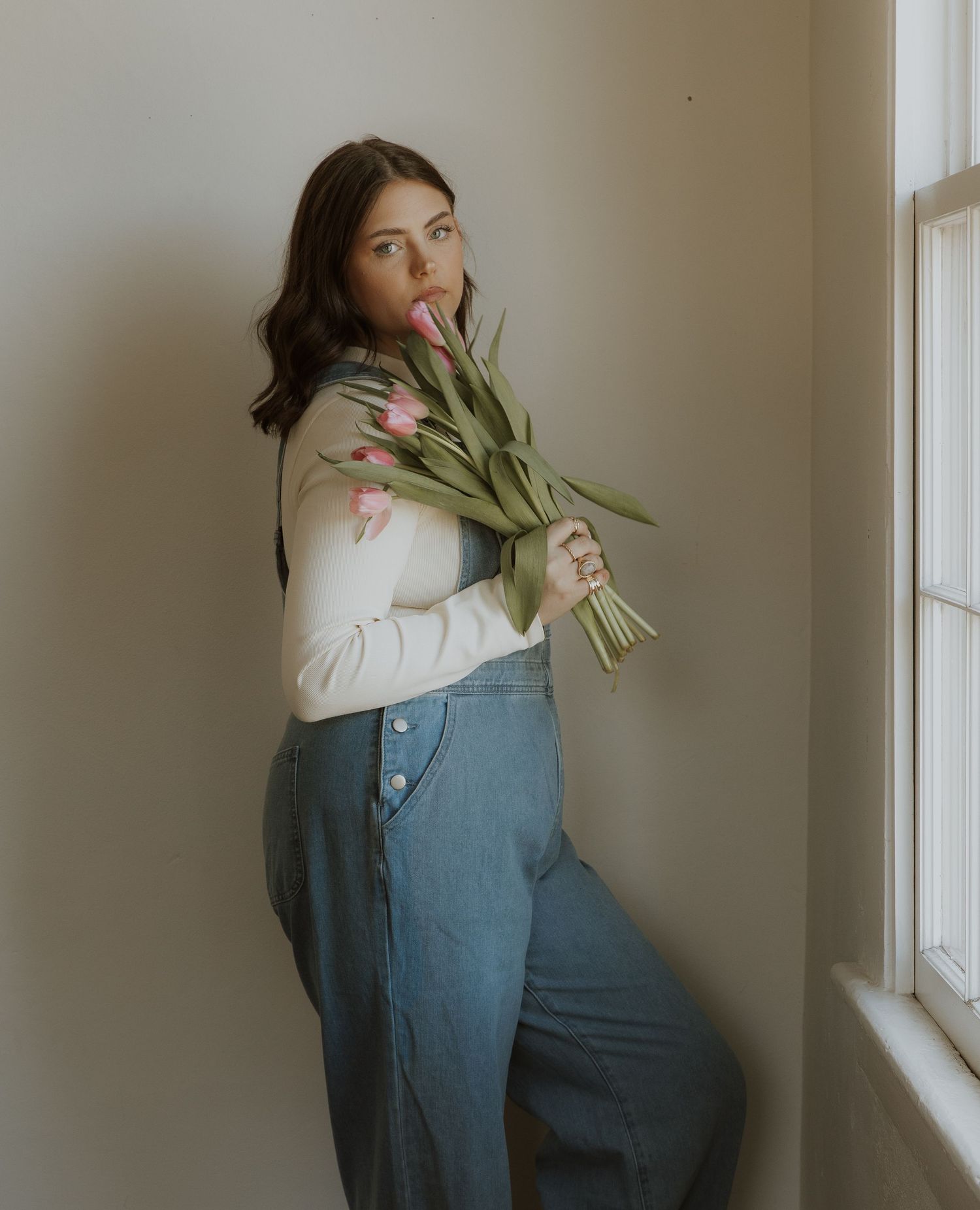 Someone in denim and a white sweater poses with white tulips by a sunny window.