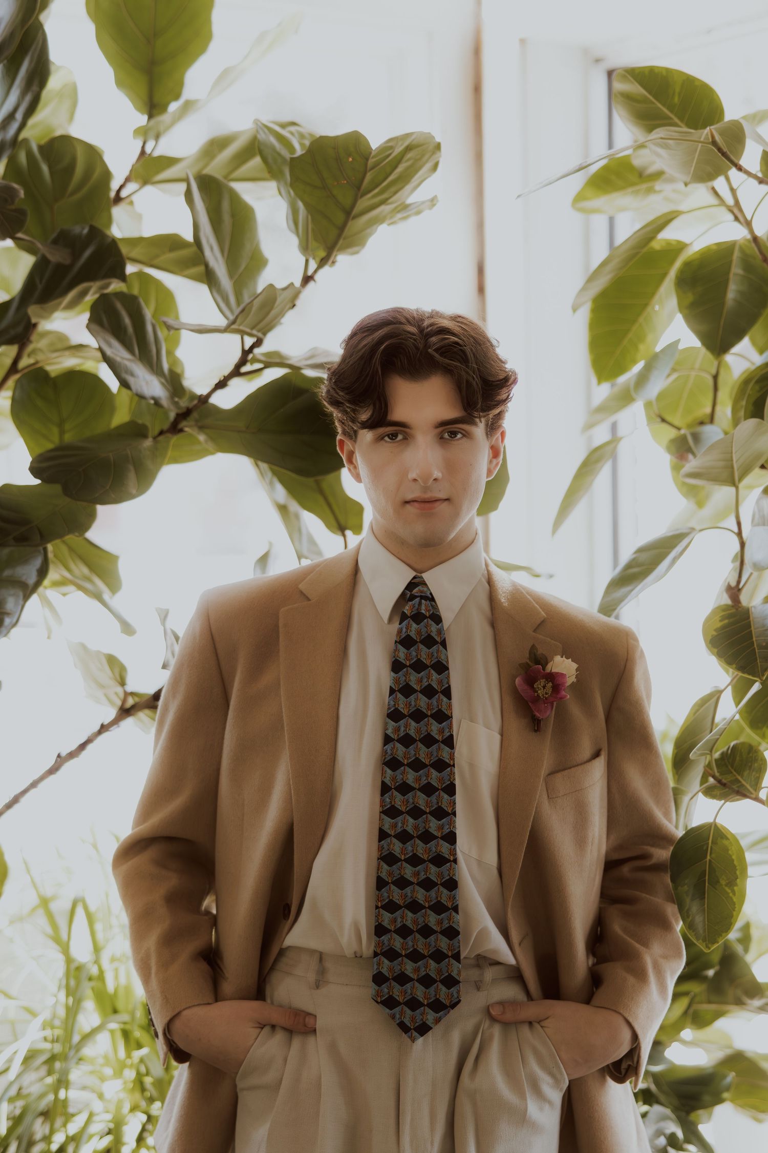 Young man in tan suit and patterned tie posed among green foliage.