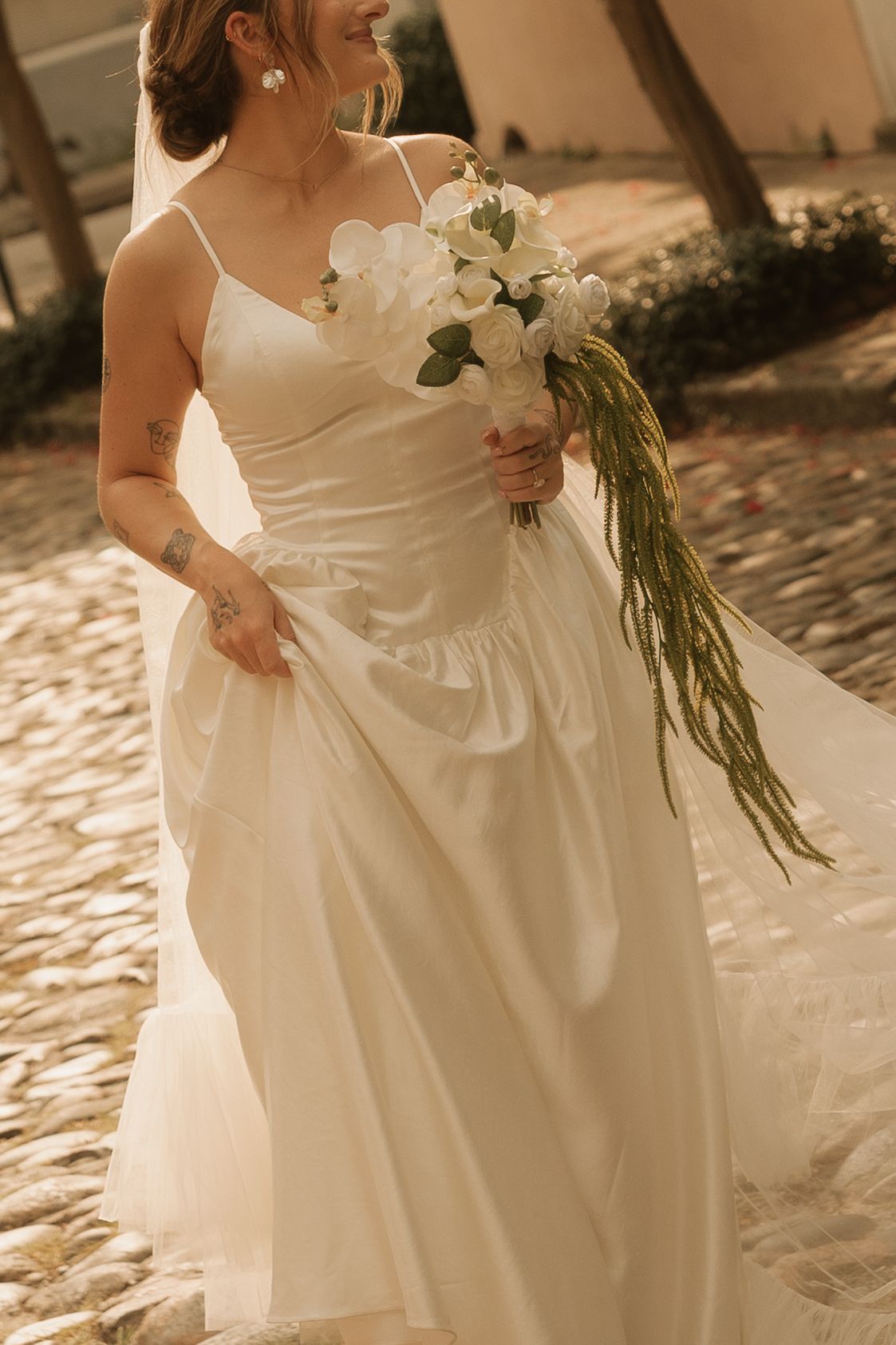 Bride in elegant white spaghetti strap wedding gown holding white floral bouquet on cobblestone path.