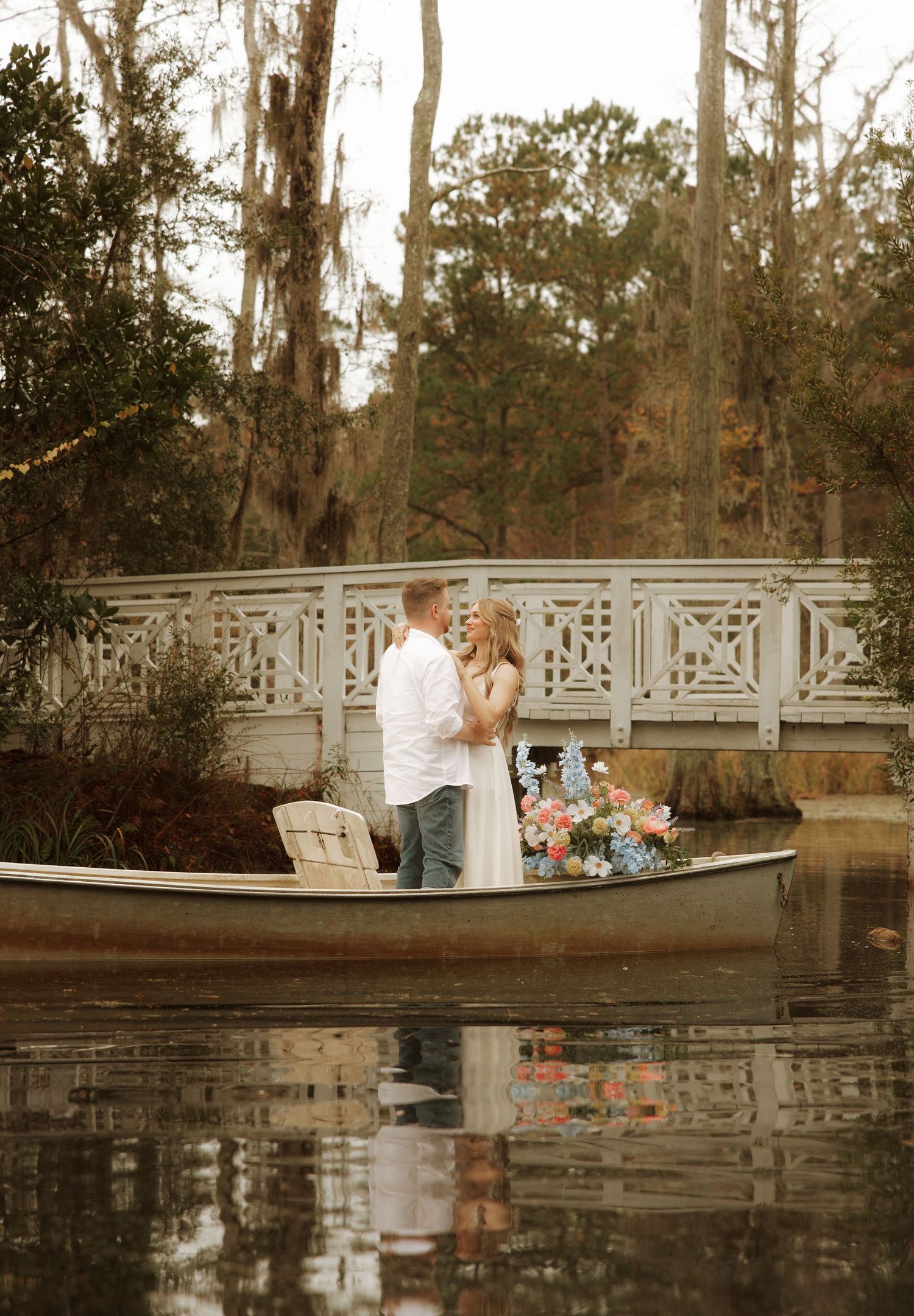 Couple standing in a canoe on a reflective lake near a white ornate bridge, surrounded by trees and colorful flowers.