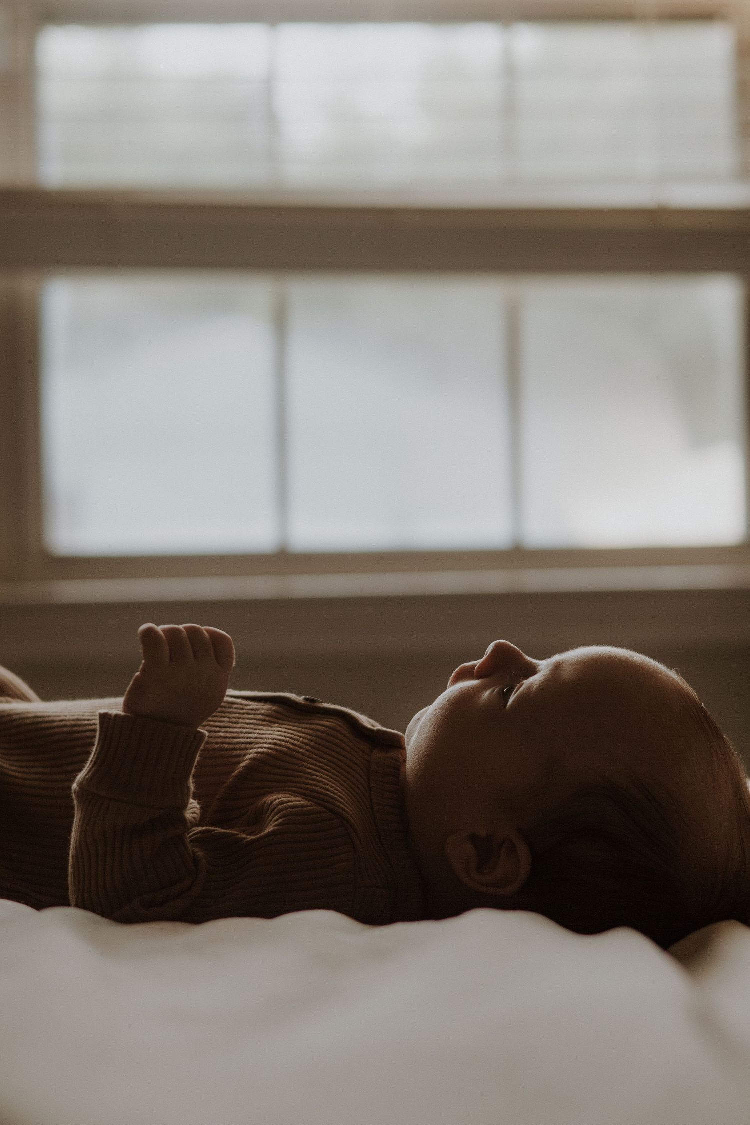 Infant in brown sweater resting peacefully on white bedding in moody lighting.