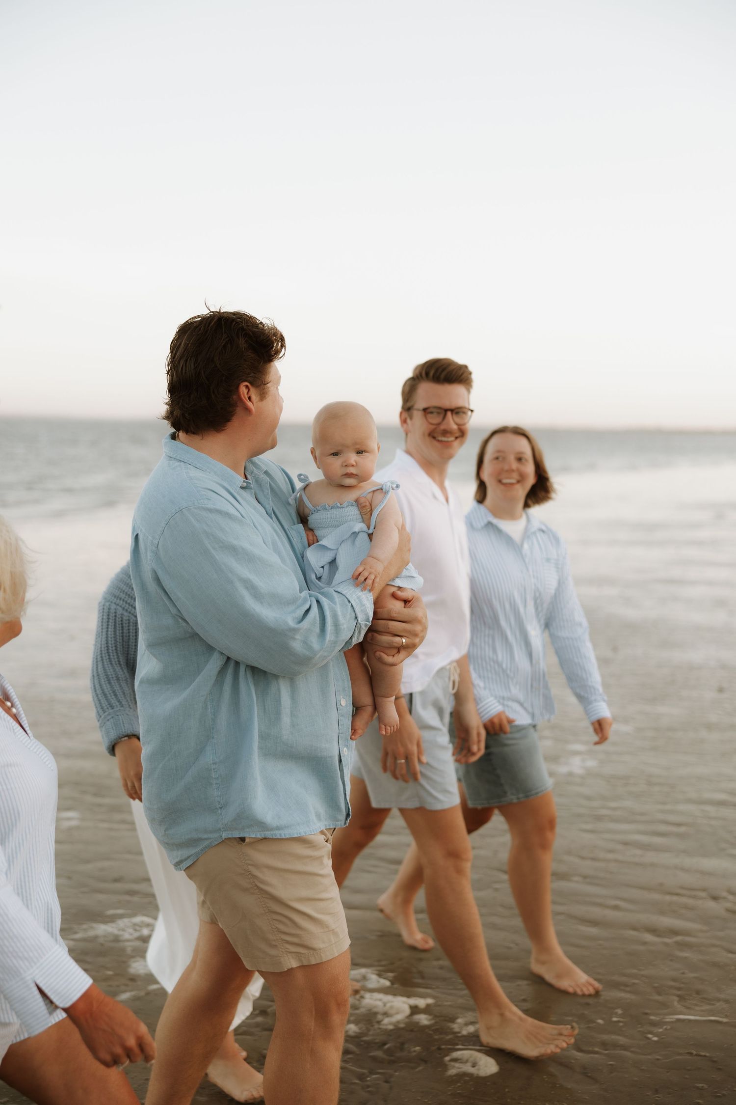 A family enjoys a casual stroll along the beach with their baby during sunset, wearing coordinated blue and white outfits.