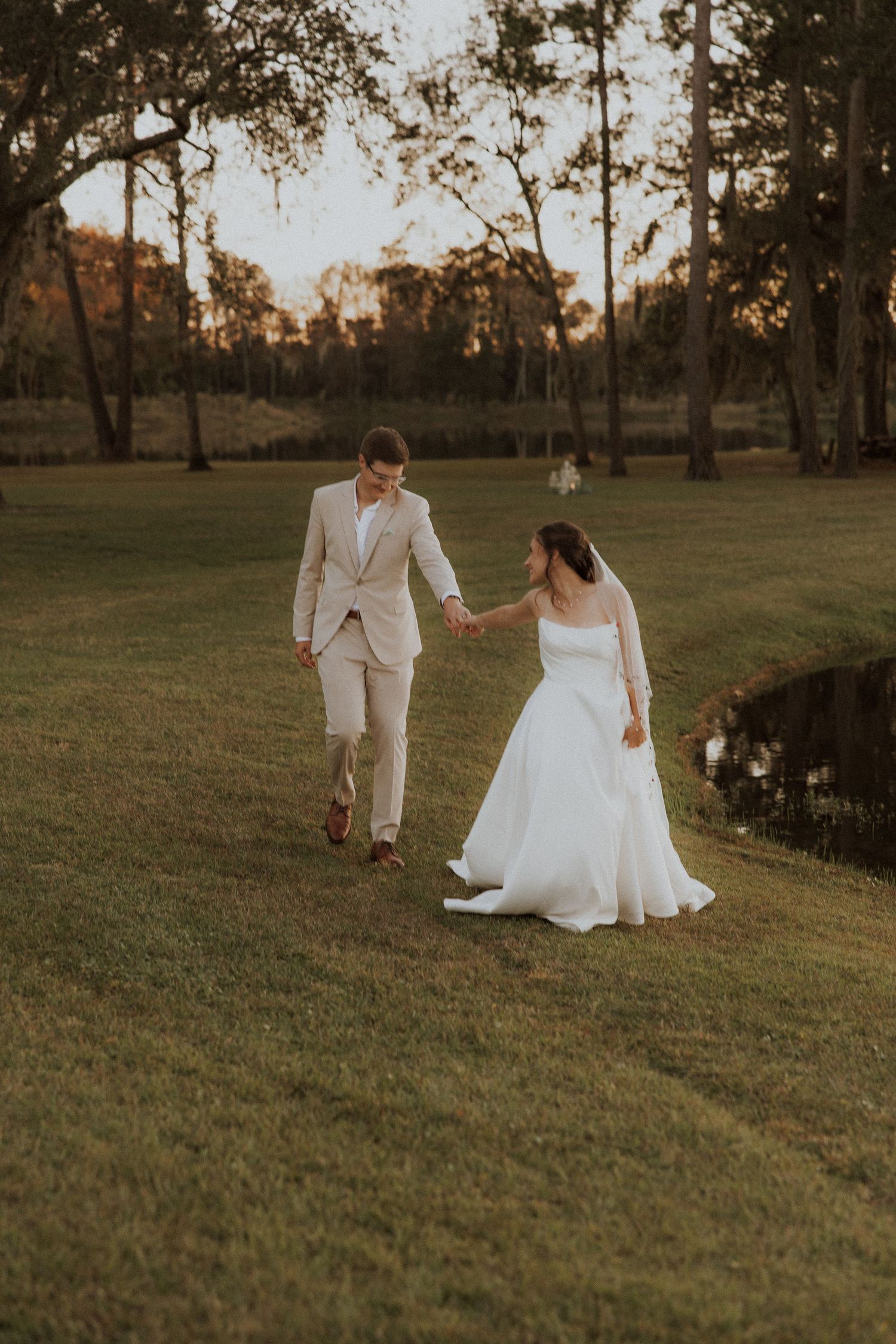 Newlyweds walking hand in hand during golden hour beside a pond with trees in the background.