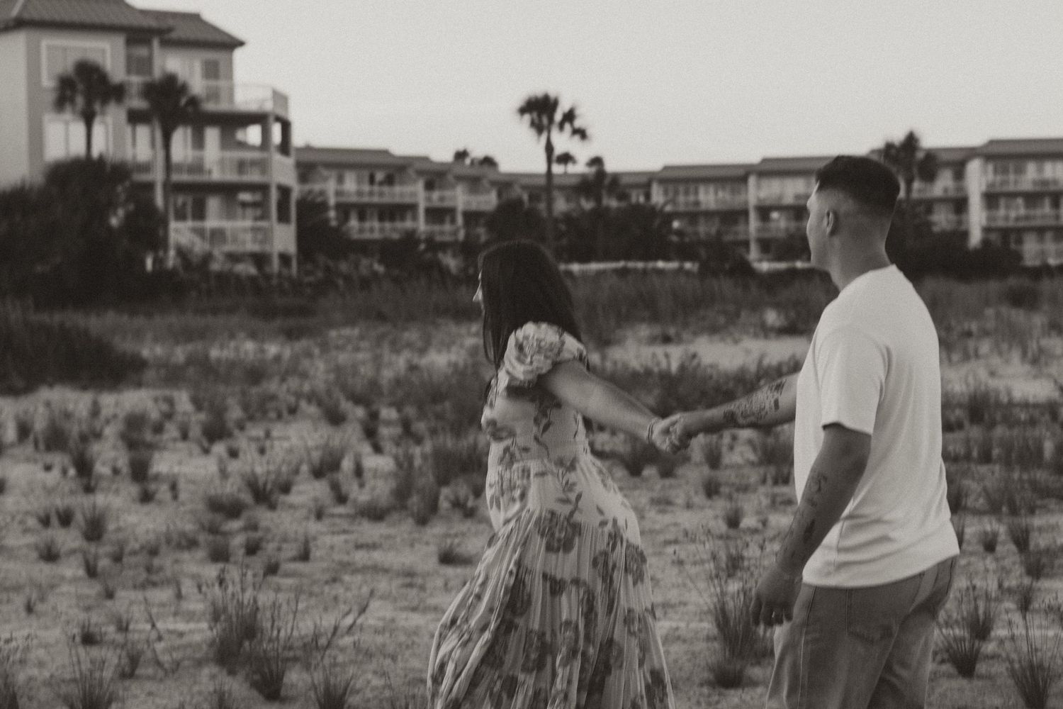 A couple holding hands on a beach with condominiums in the background.