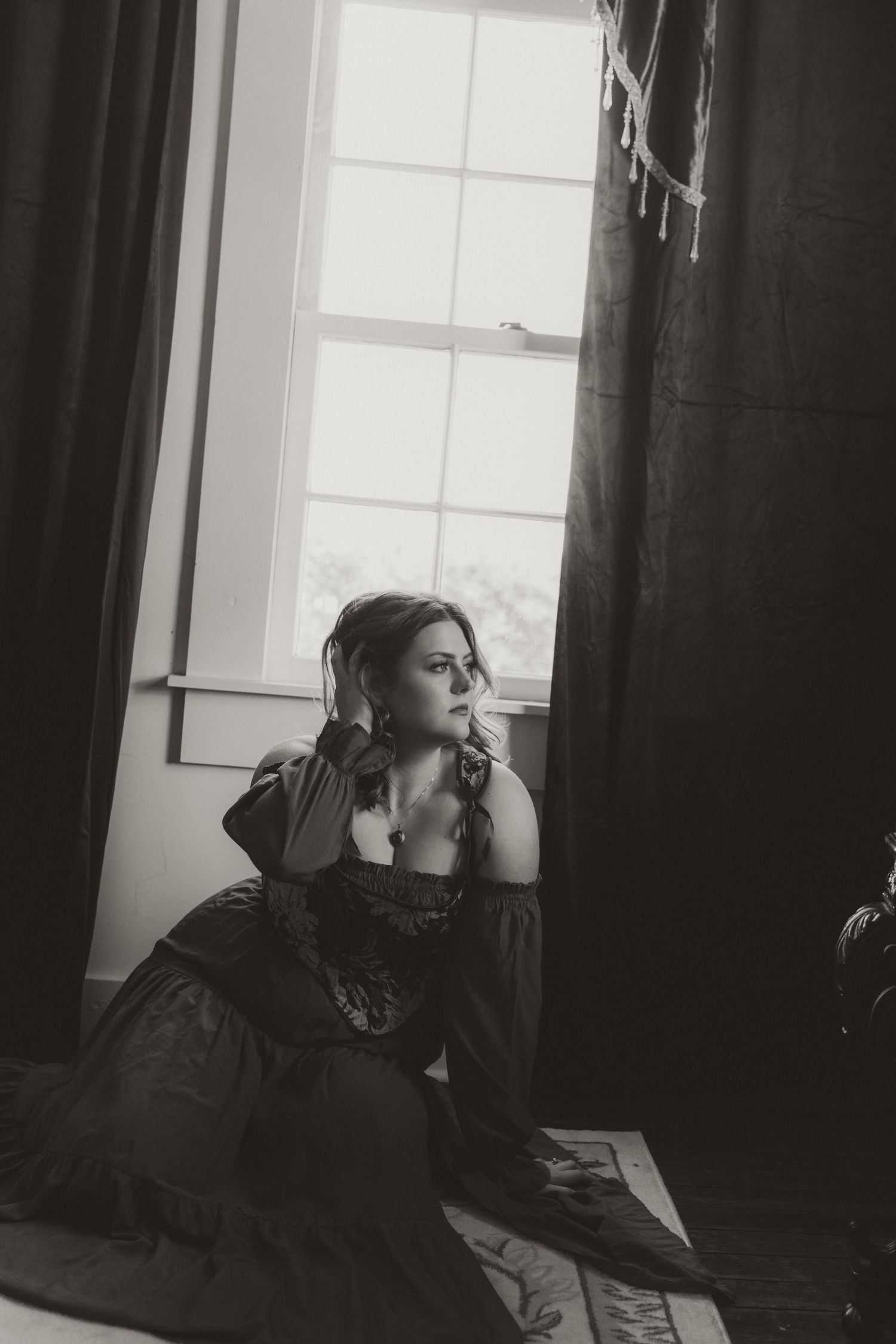 Black and white photo of a woman in a Victorian dress sitting by a window with dark curtains, gazing thoughtfully.
