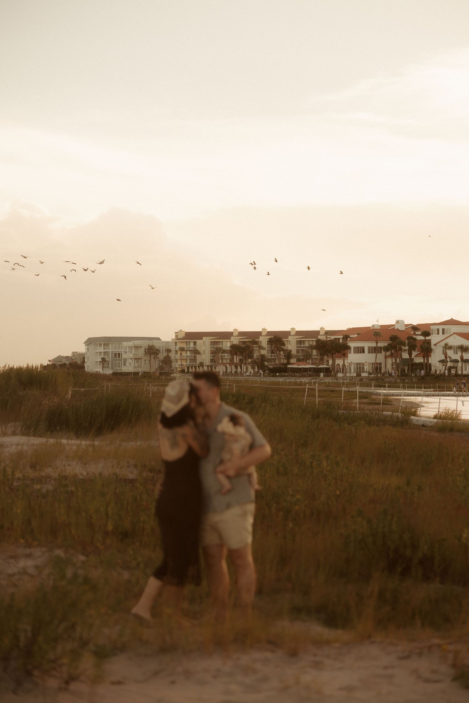 Couple embracing on a sandy beach at sunset with coastal buildings and birds flying in the warm evening sky.