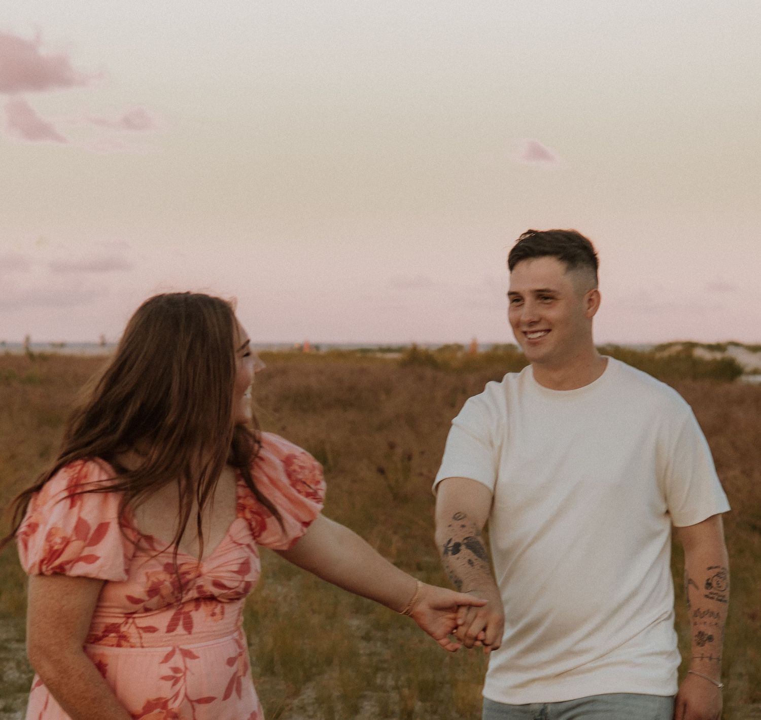 Couple holding hands in field at sunset wearing pink floral dress and white t-shirt.