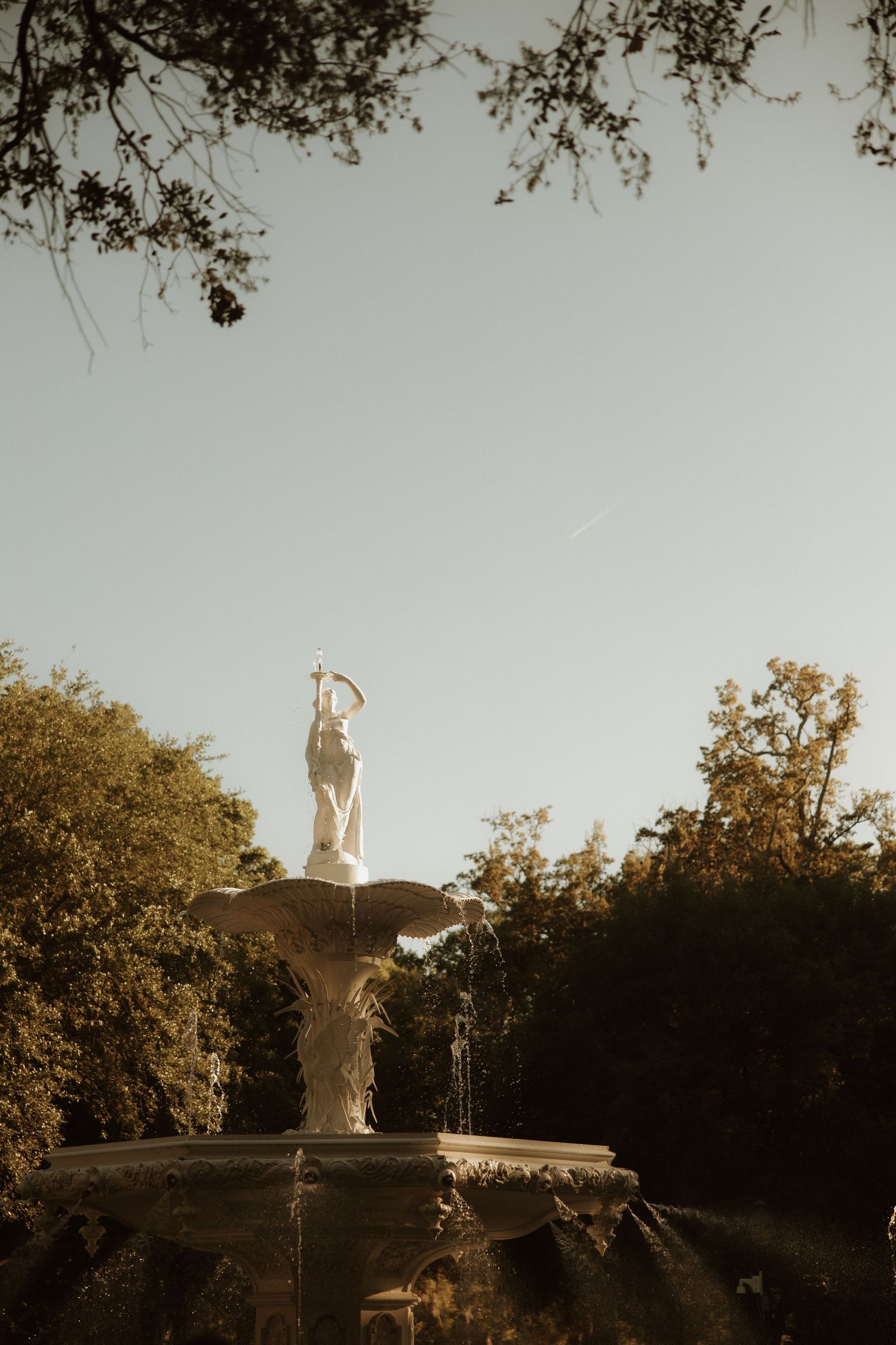 White marble fountain statue surrounded by lush green trees against a clear sky in golden hour light.