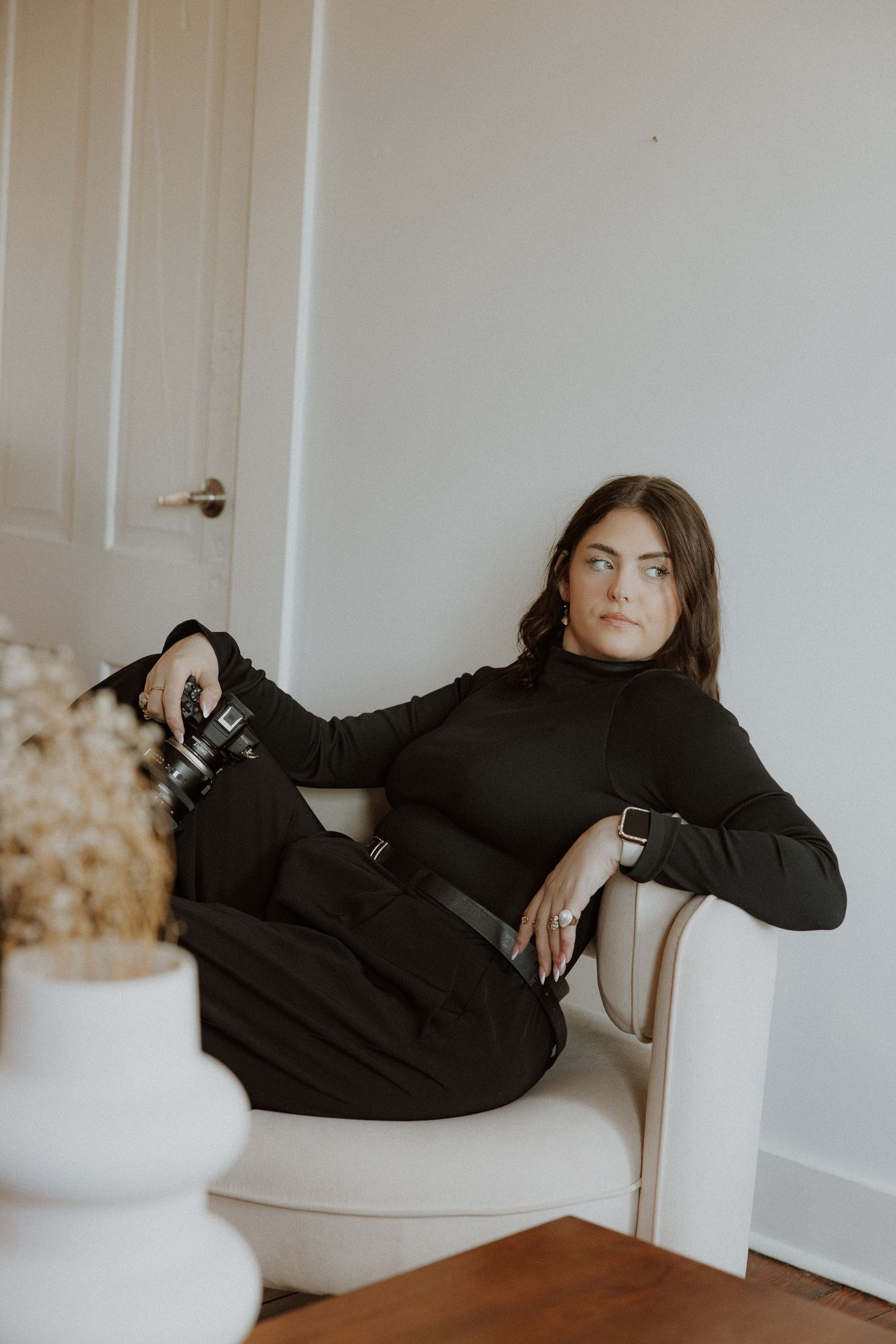 Person in black turtleneck sits elegantly in white chair with camera nearby.