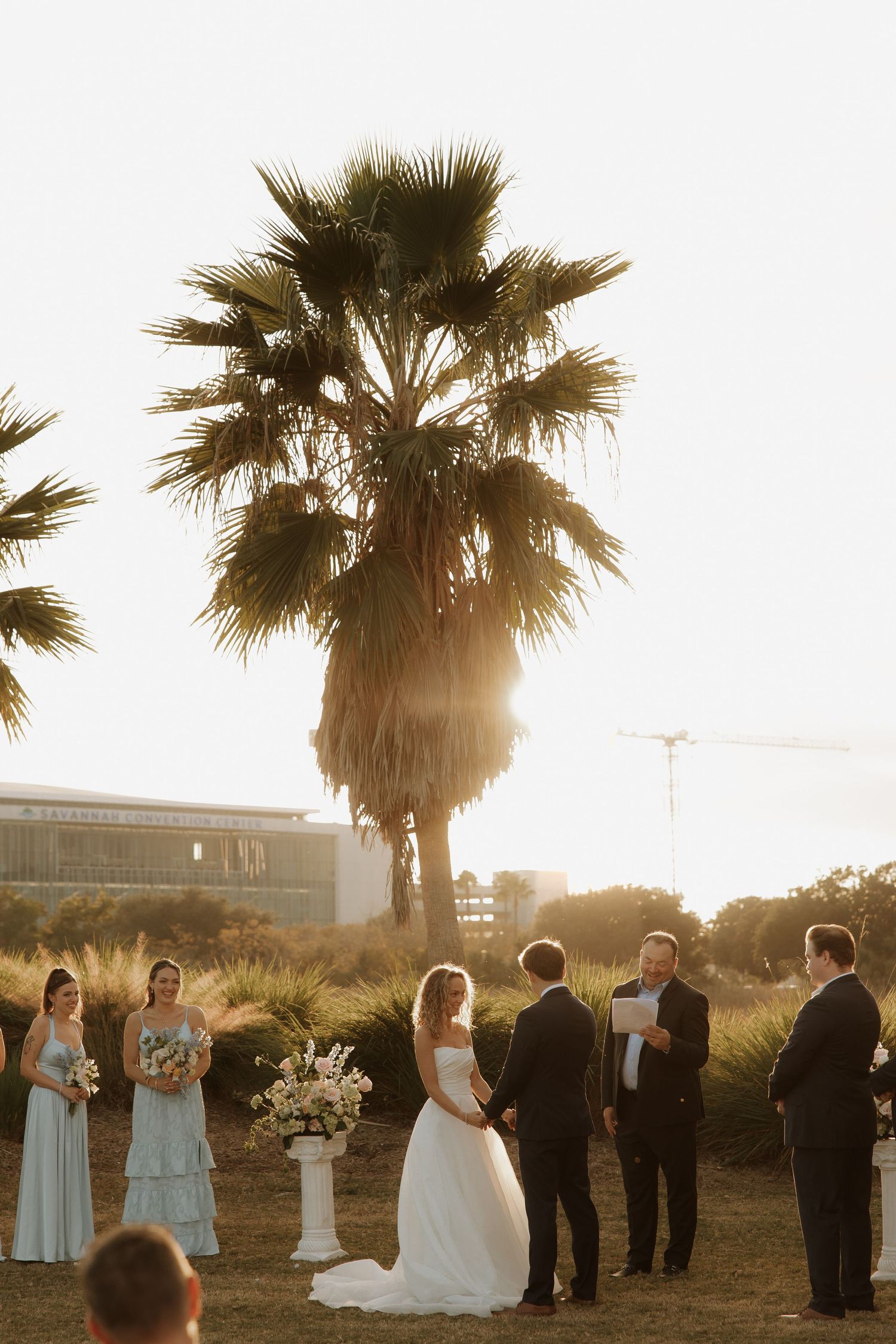Wedding party stands in line under palm trees during golden hour with sunlight streaming through.