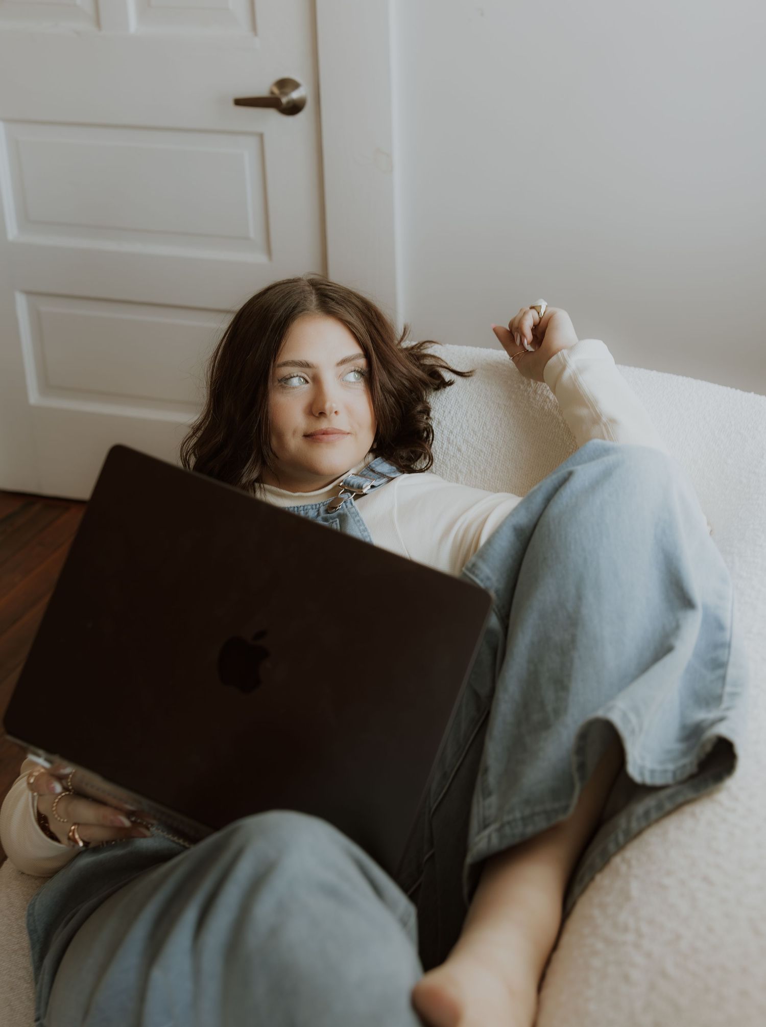 Person relaxes on white couch with MacBook laptop wearing denim overalls.