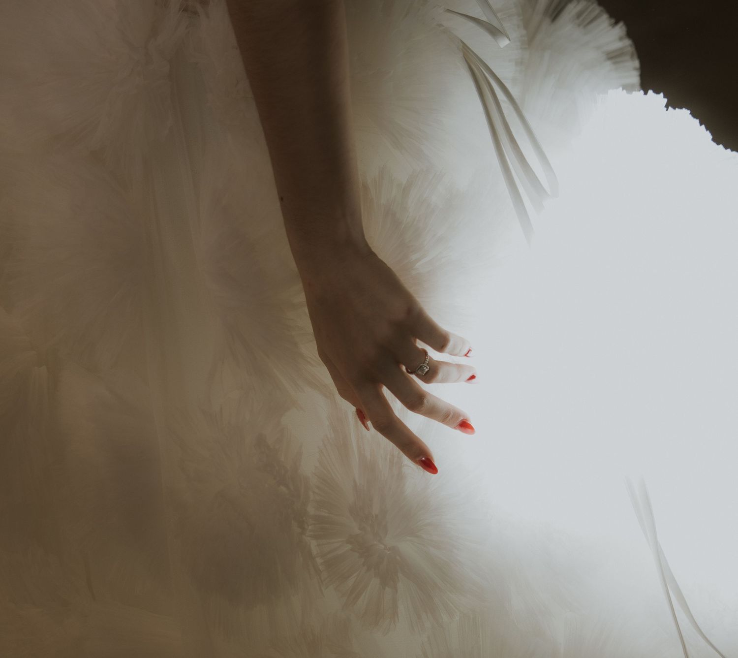 Close-up of a hand with engagement ring against white fabric in dreamy lighting.