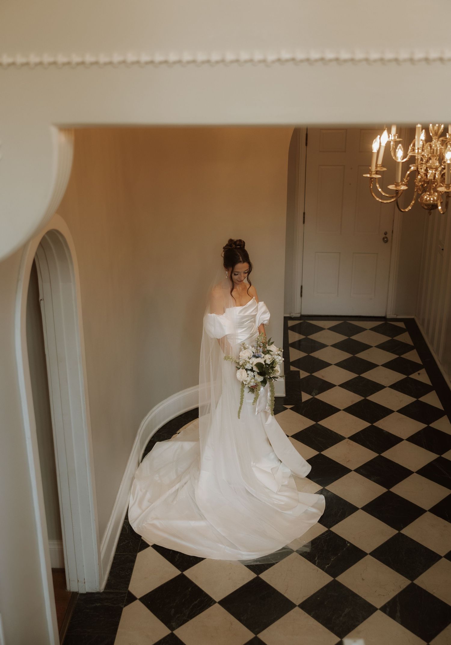 A bride in a flowing white gown twirls on a black and white checkered floor in an elegant room with arched doorways.