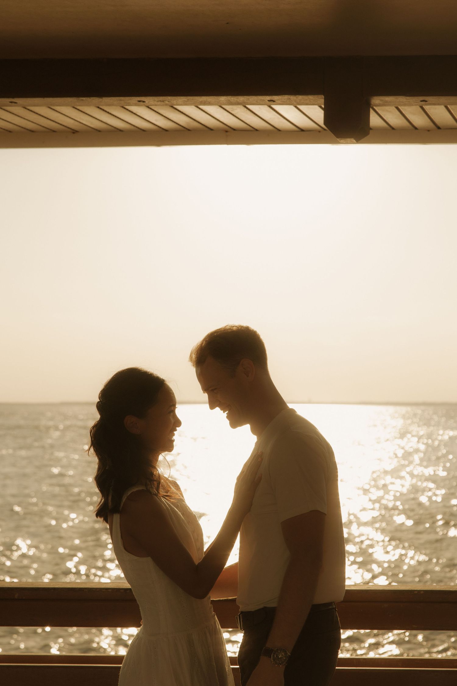 Romantic couple holding hands facing each other at sunset by the ocean on a waterfront deck.
