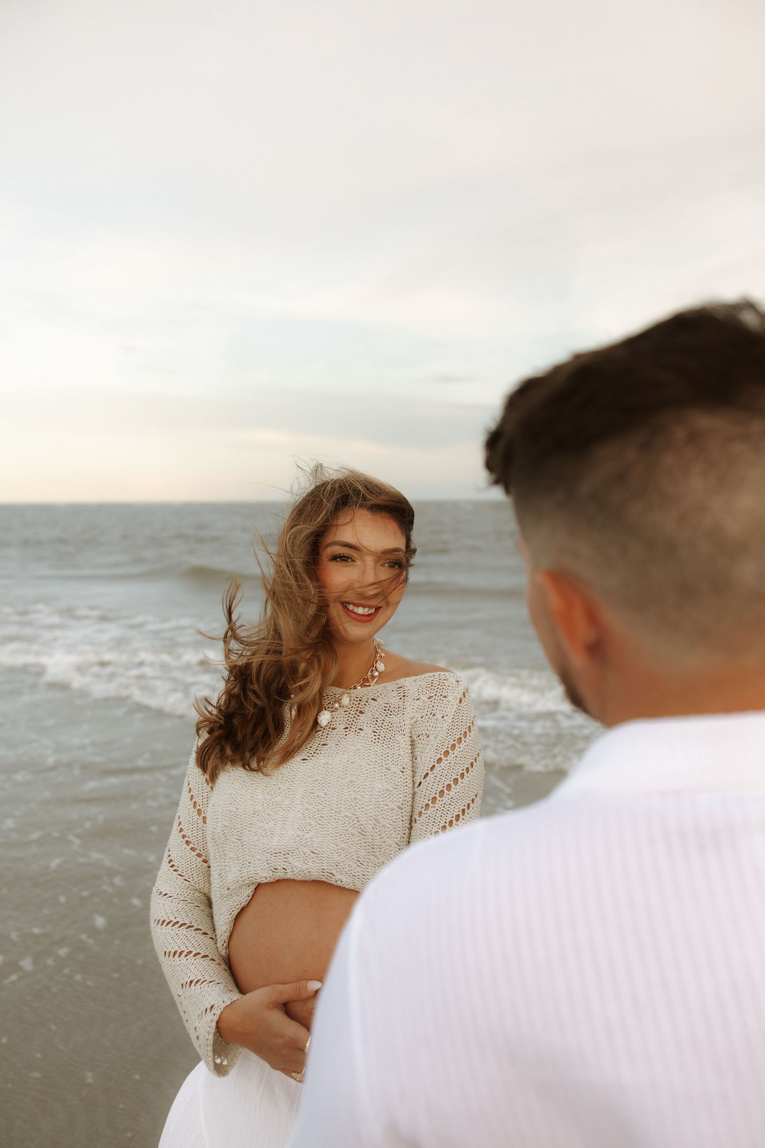 Romantic coastal photo session featuring a couple in white outfits posing together on a beach at sunset.