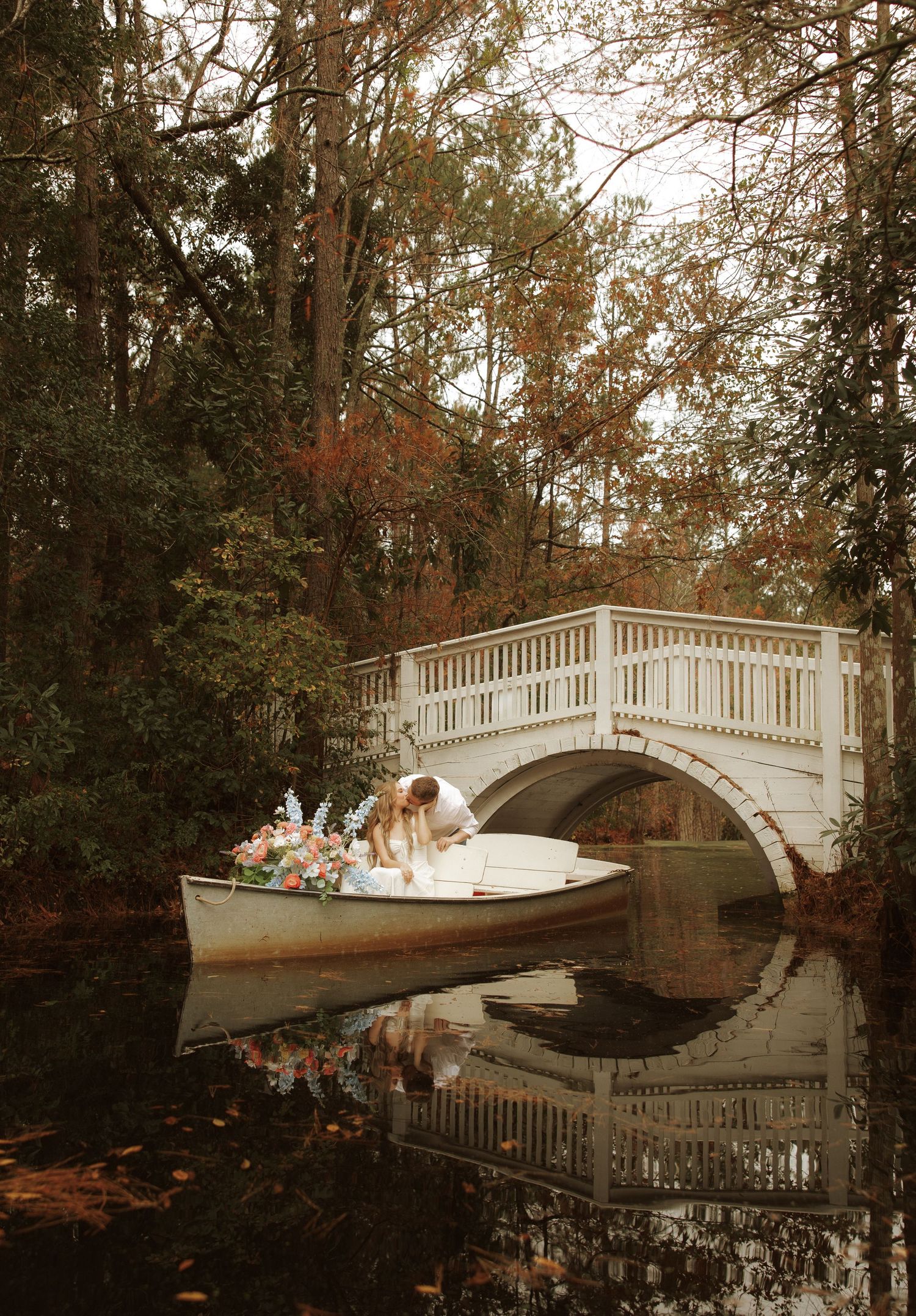 A romantic boat scene with a white arched bridge over calm reflective water in an autumn woodland setting.