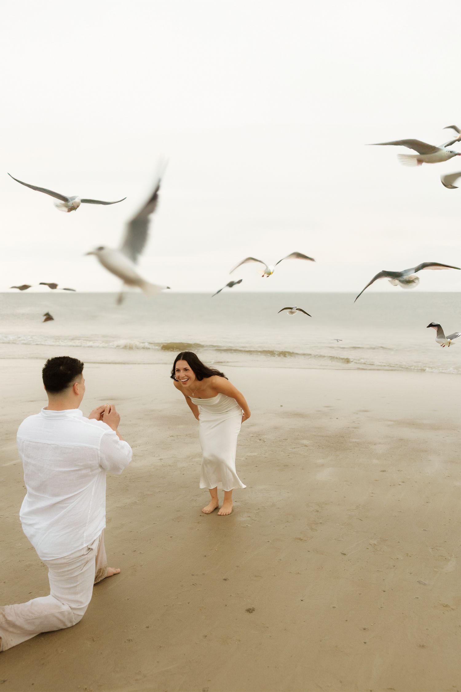 Couple in white outfits taking photos on a beach with seagulls flying overhead during a romantic photoshoot.