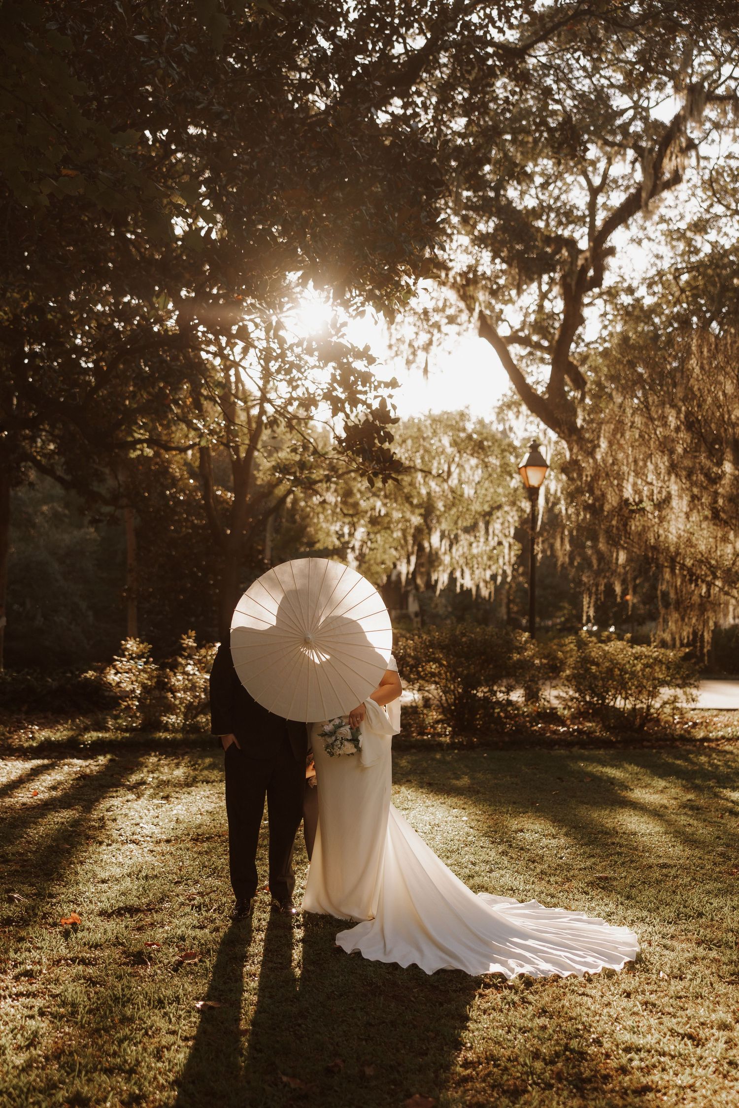 Silhouetted couple embraces in golden sunlight filtering through Spanish moss-draped trees during sunset wedding ceremony.