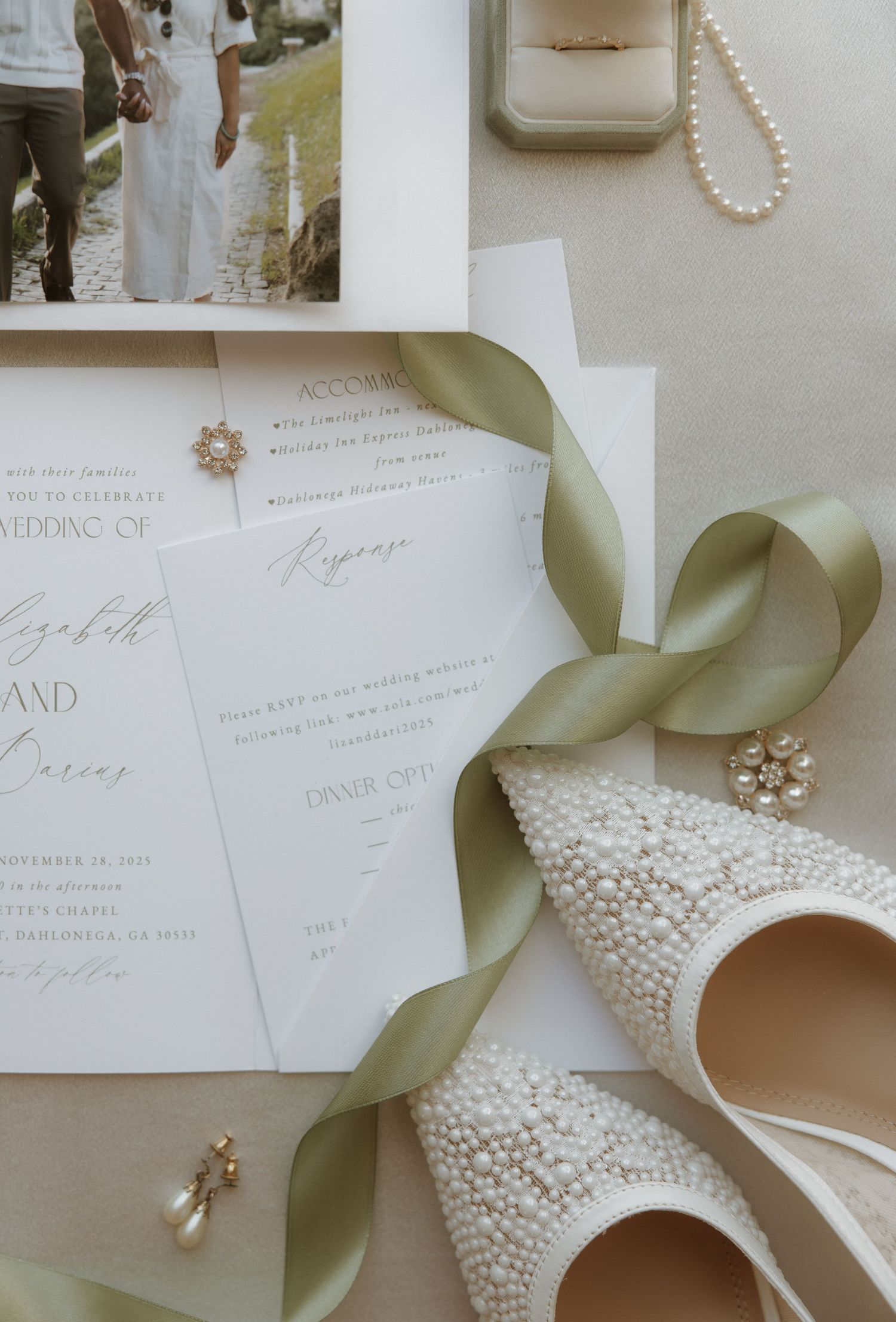 Elegant wedding invitation suite with pearl details, sage green ribbon, and crystal embellishments laid out on white surface.