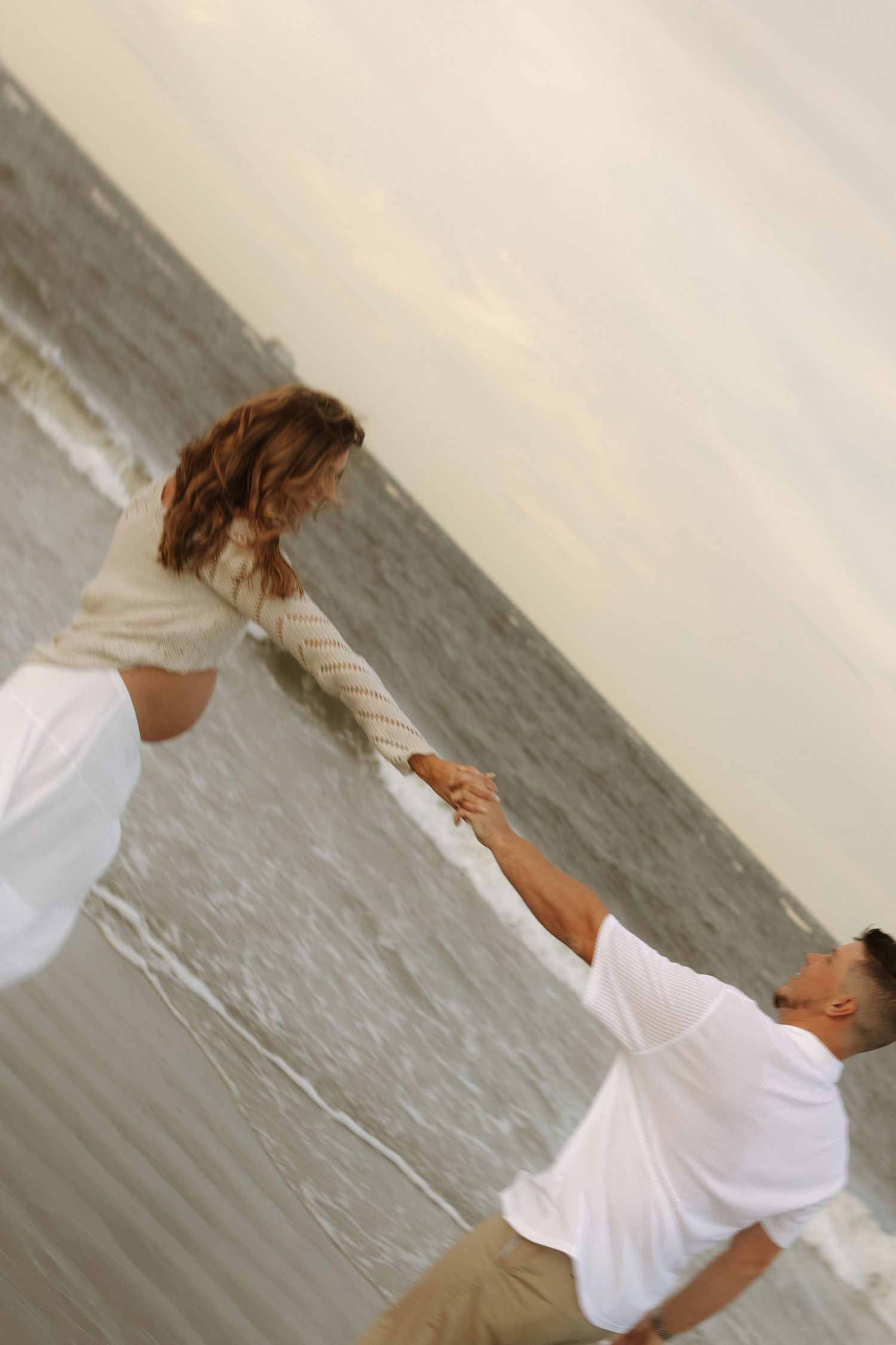 A couple in white clothing dances gracefully together on a beach with water in the background.