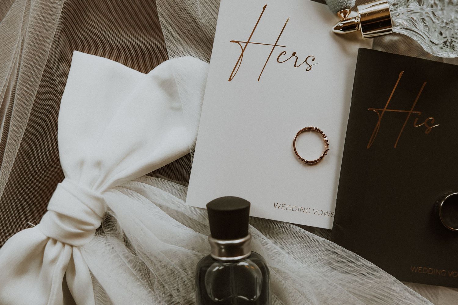 Wedding details including vows book, white tulle fabric, and gold wedding ring on neutral background.