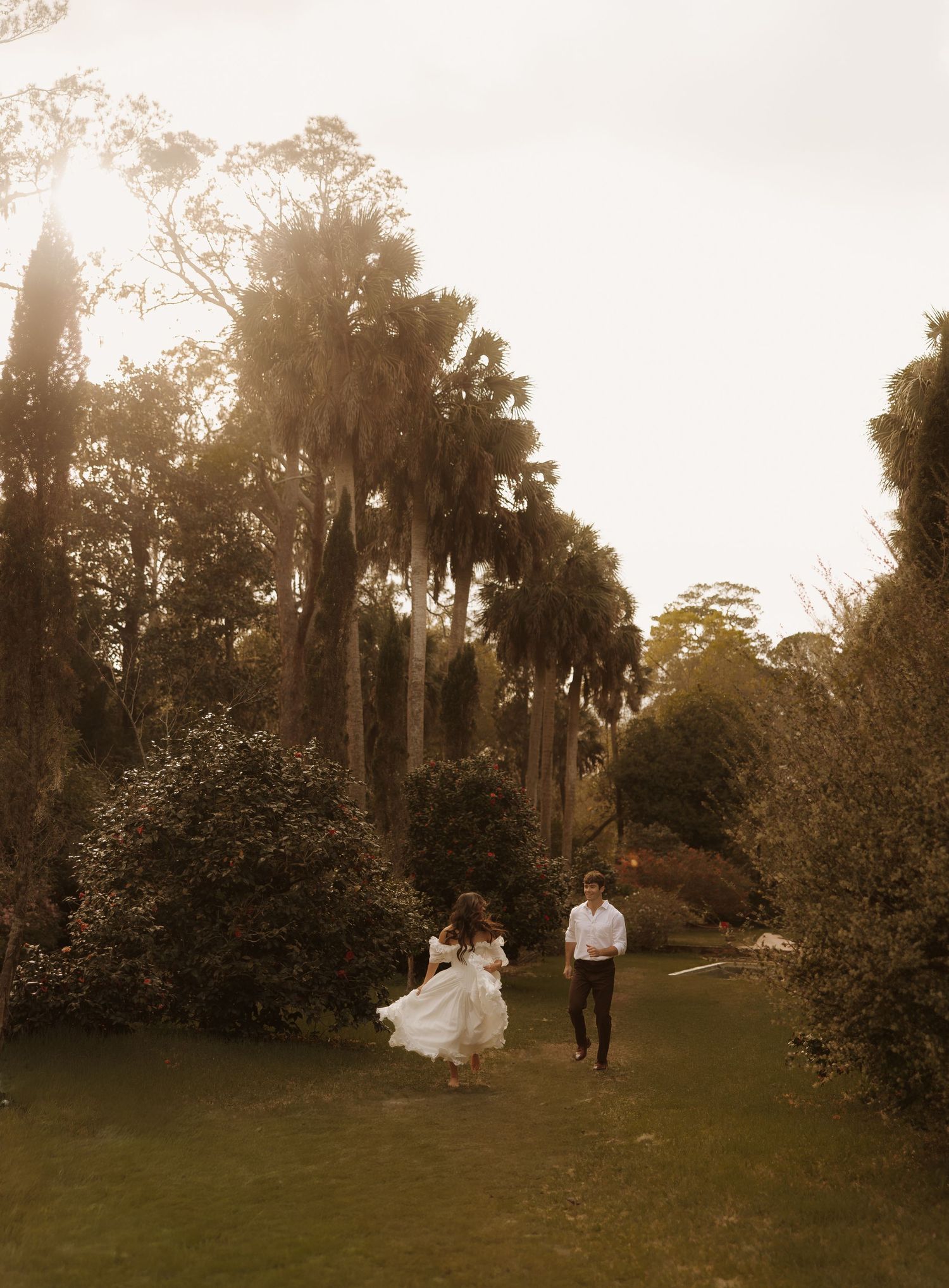 Bride and groom running through a sunlit garden with tall palm trees during golden hour wedding photography.