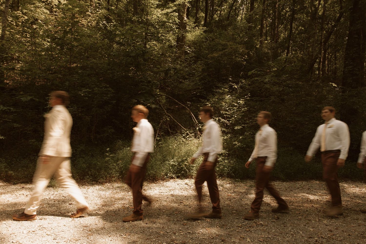 Blurred motion of men in formal attire walking along a sunlit forest path surrounded by lush green trees.