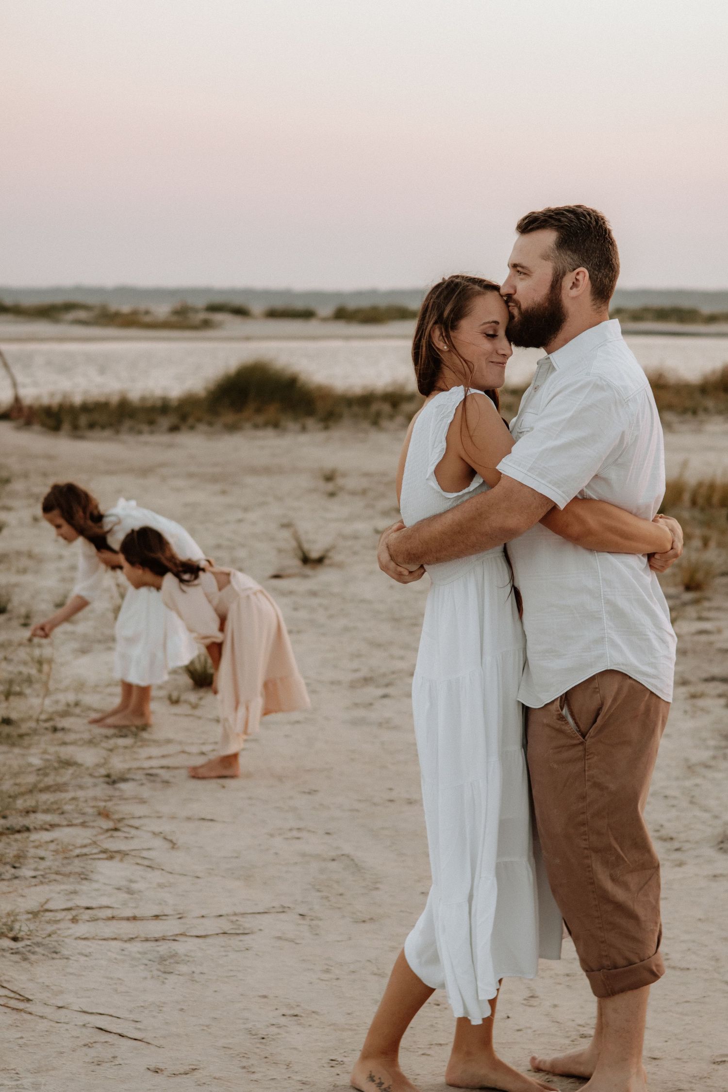 A couple embraces on the beach while others play in the background during sunset.
