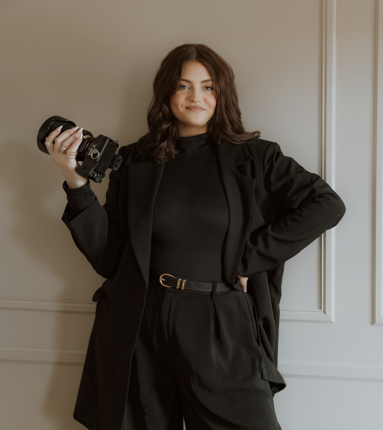 Photographer in black outfit with camera against neutral wall interior.