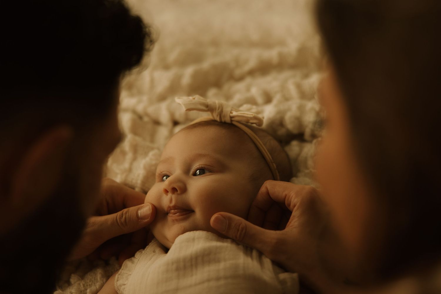Newborn baby girl wearing a bow headband lying on white blanket while parents gently touch her cheeks.