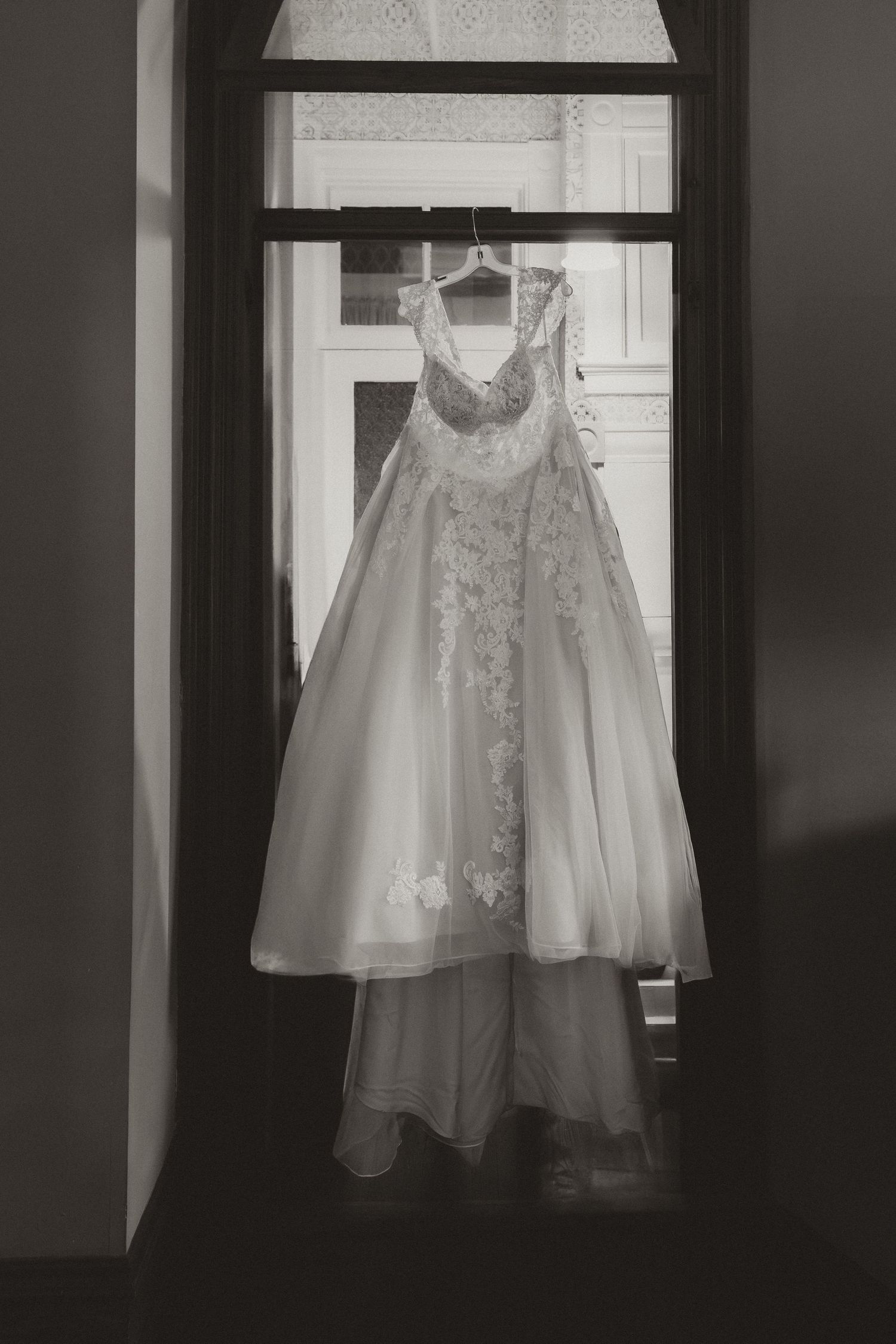 Elegant white lace wedding dress hanging in a doorway, captured in black and white.