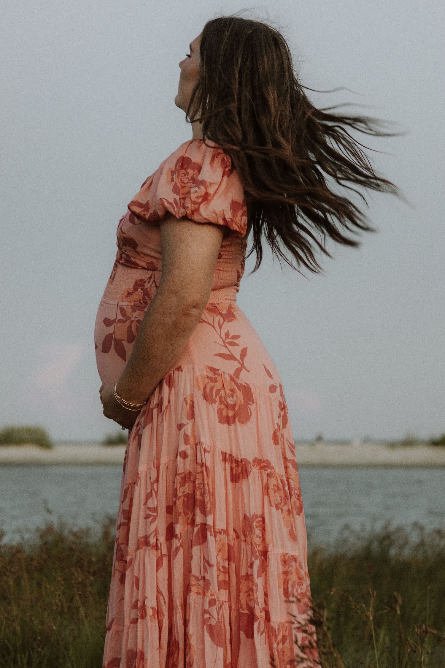 Woman in pink floral dress stands by water with hair blowing in wind.