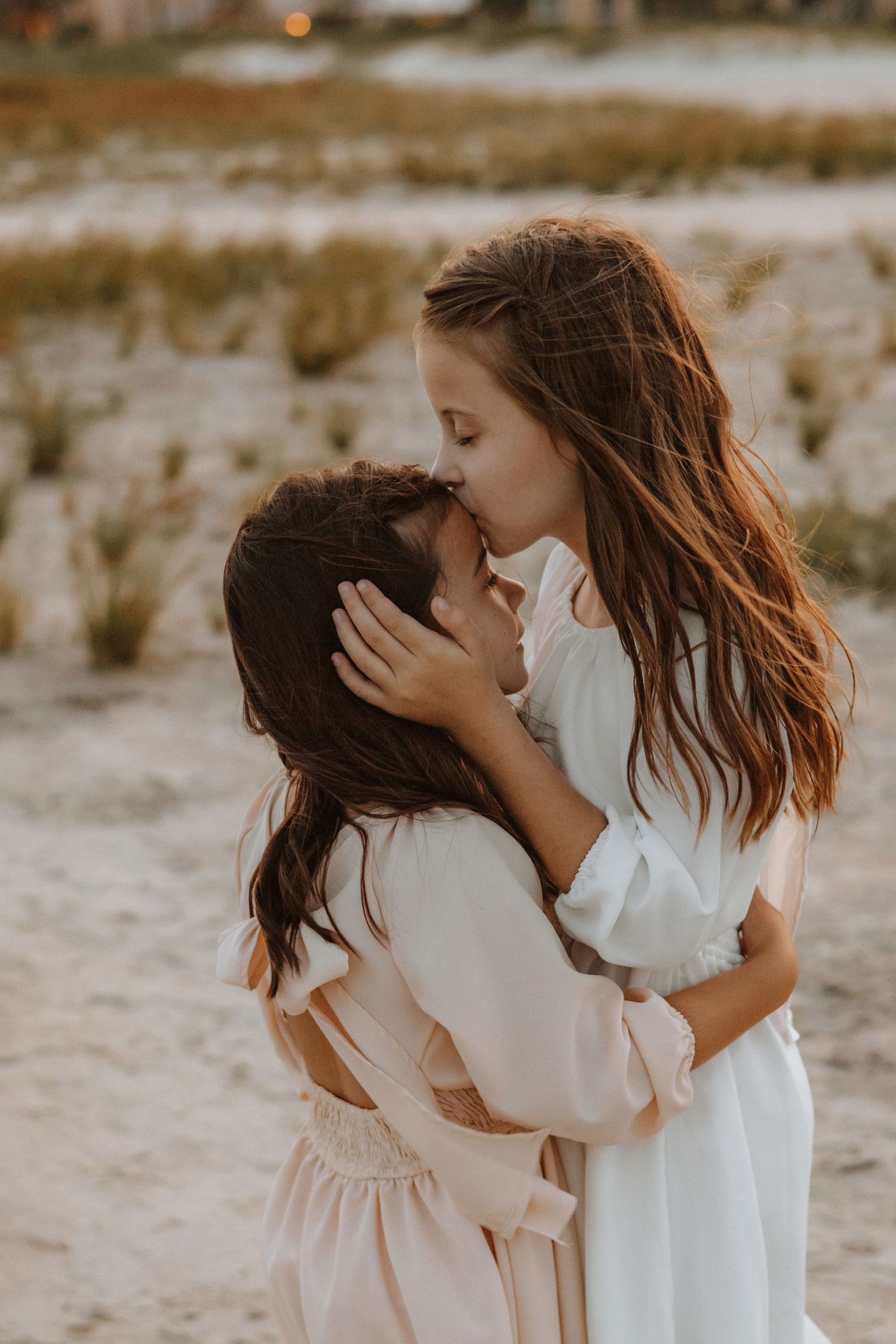 Two young children in white dresses sharing a tender moment outdoors.