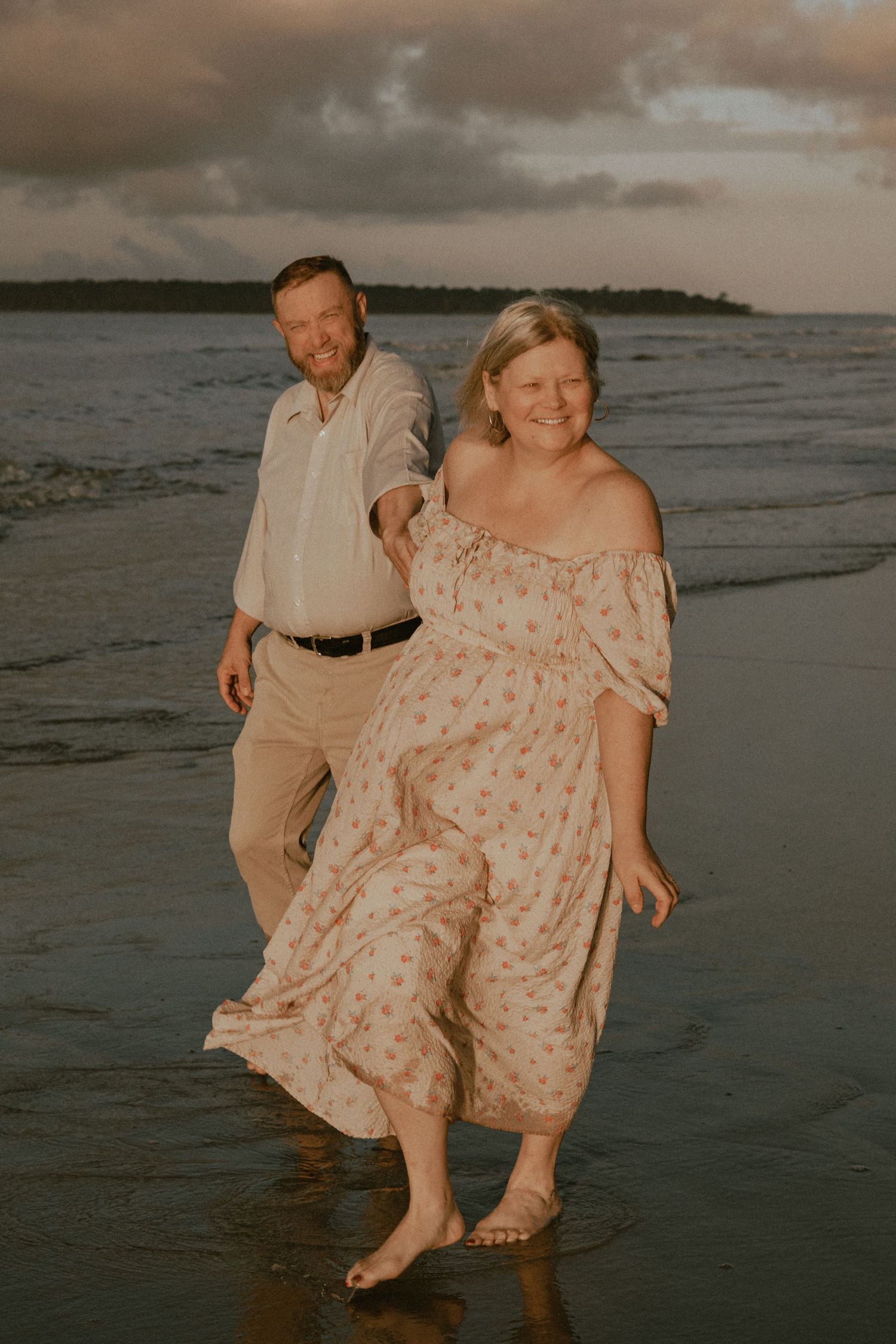 Couple walking barefoot in the shallow water along the shoreline at dusk.