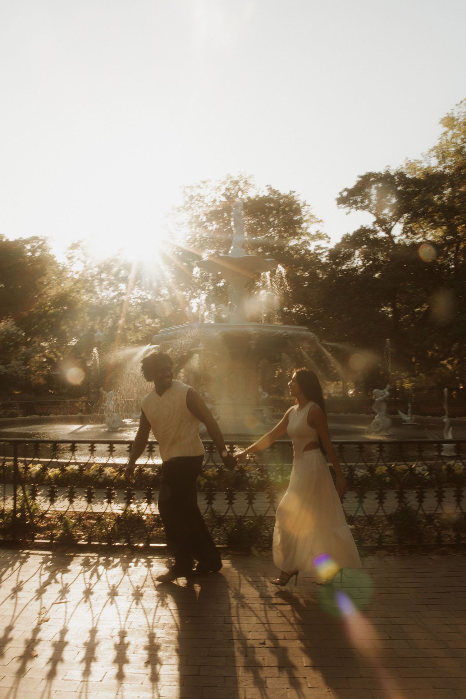 Couple holding hands walking by a sunlit fountain in a park, golden hour light with lens flare, woman in white dress.