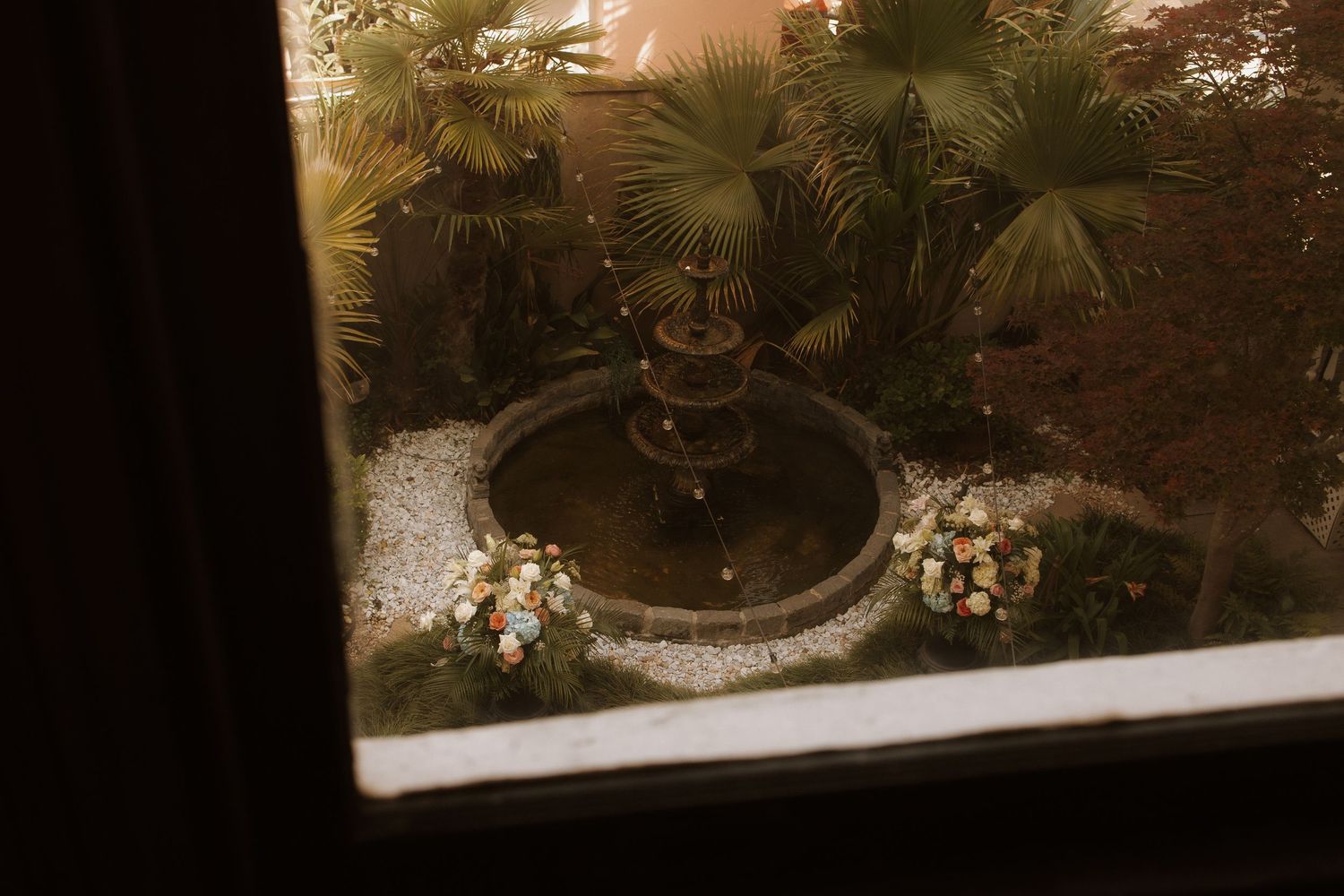 A tiered stone fountain surrounded by tropical palm trees and colorful flowers, viewed through a window at night.