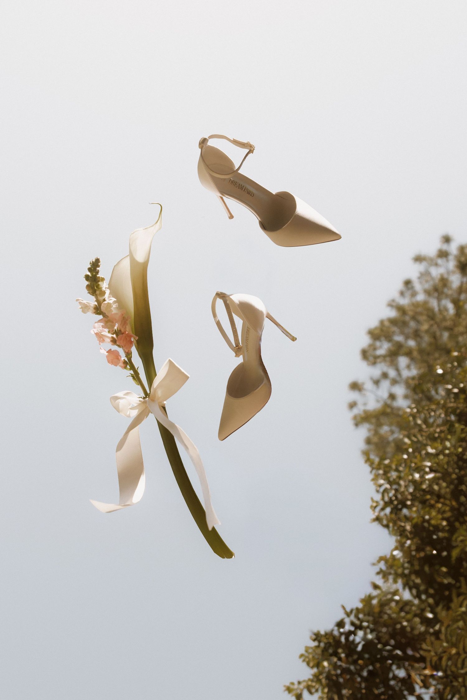 Elegant nude satin heels and floral bouquet with ribbon floating against a soft sky background.